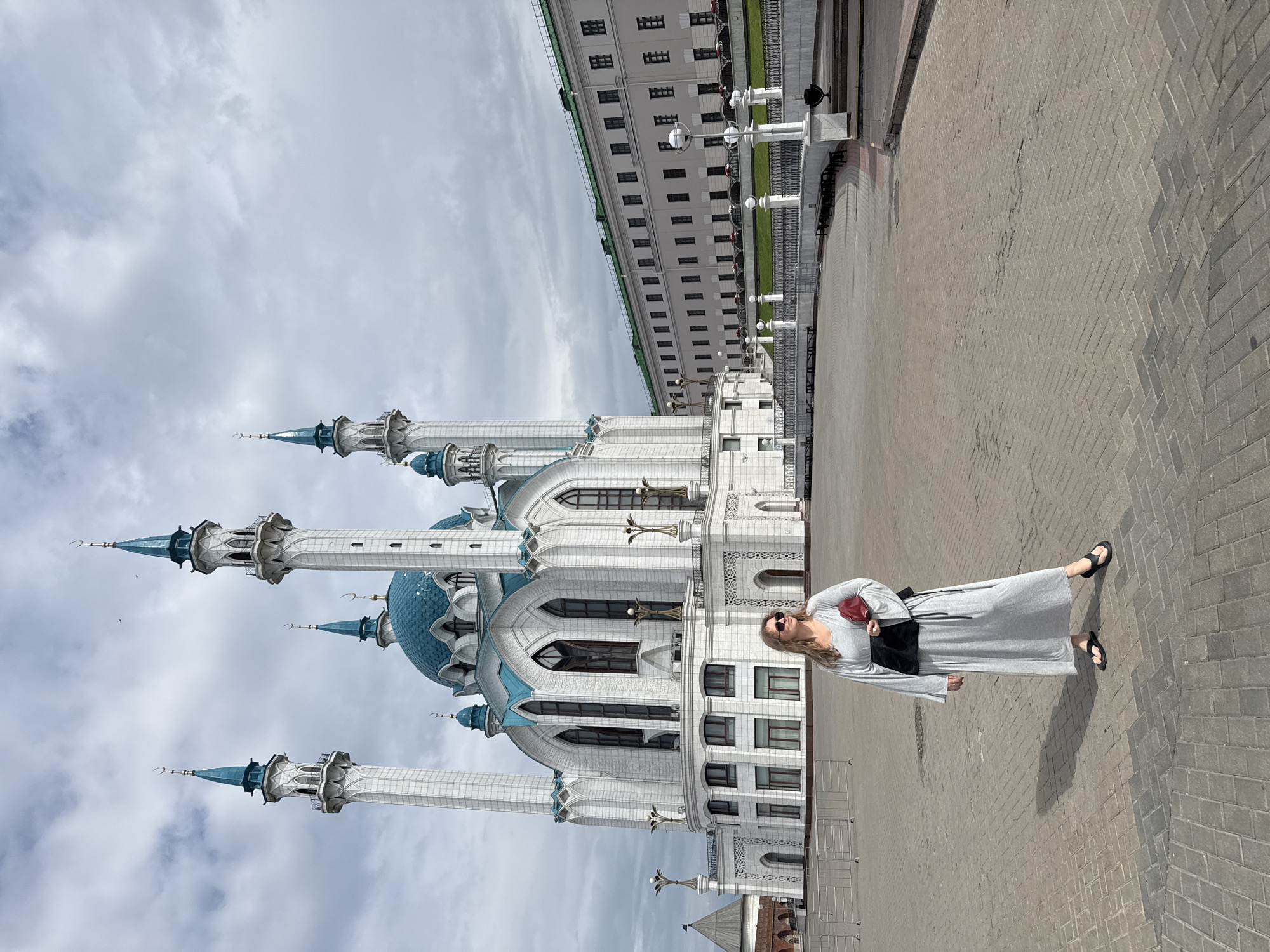 A woman stands in a paved plaza in front of the Kul Sharif Mosque in Kazan, Russia. She is wearing a long gray dress, a black sash, and black sandals. She is holding a red clutch purse. The mosque, a prominent landmark of Kazan, features white walls, blue domes, and tall minarets. To the right of the mosque, a long, multi-story building with a green roof stretches into the background. The sky is overcast with scattered clouds. The ground is paved with light-colored bricks, and several metal barriers are visible in the foreground.