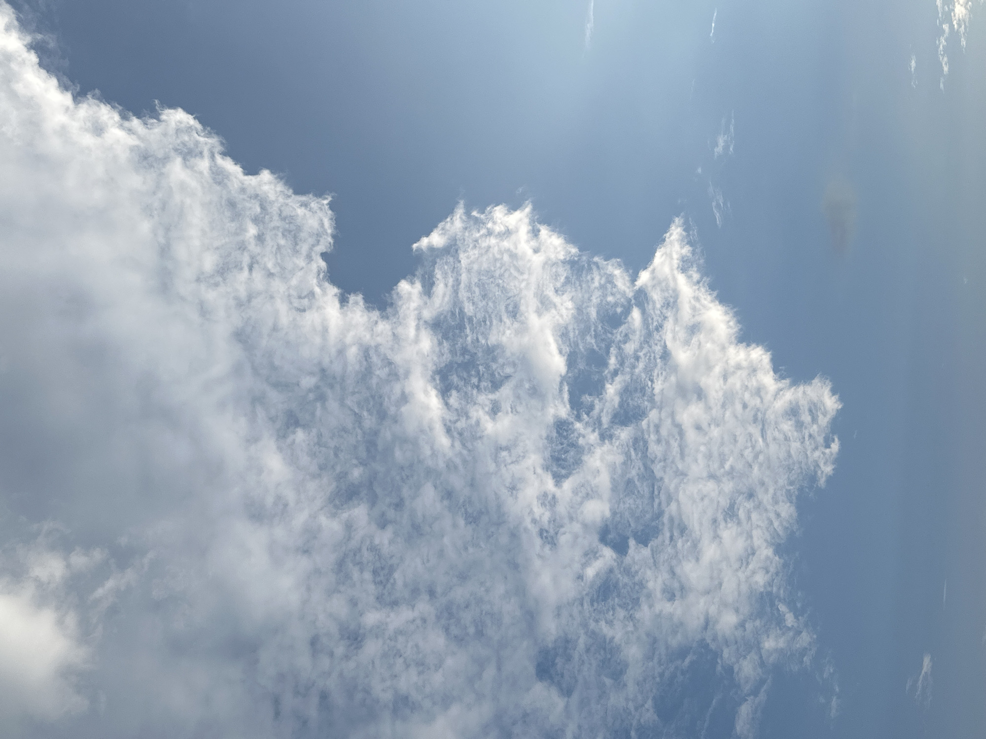 The image is a vertical shot of the sky, featuring a clear blue sky on the right side and a large, textured cloud formation on the left. The cloud appears to be a cumulus cloud with a bright, white, fluffy top and a more wispy, scattered base. The lighting suggests it is daytime, with the sun likely positioned behind or to the side of the clouds, illuminating them from above. There are no people, buildings, or any other objects visible in the frame. The scene is entirely focused on the natural elements of the sky. There is no identifiable location within the city of Chaikovsky, Russia, as the image only depicts the sky.