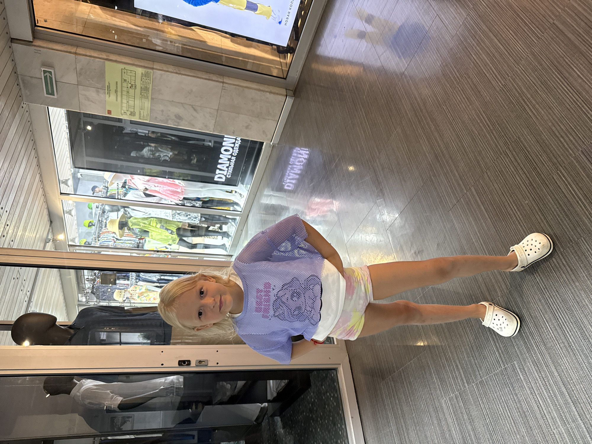 A young blonde girl stands in front of a store window. She is wearing a light purple mesh top over a white t-shirt and tie-dye shorts. She also has on white Crocs. The store window displays clothing on mannequins, with racks of apparel visible behind them. The store's name, "DIAMOND," is visible in large letters on the window. The girl is smiling and looking at the camera. The floor is tiled with a wood-grain pattern, reflecting the overhead lights. The scene appears to be in an indoor shopping area. The girl's pose and expression suggest she is posing for a photo. The lighting is bright, indicating it could be daytime. There is a sign with a green arrow pointing to the left mounted on a tiled wall above the store window.