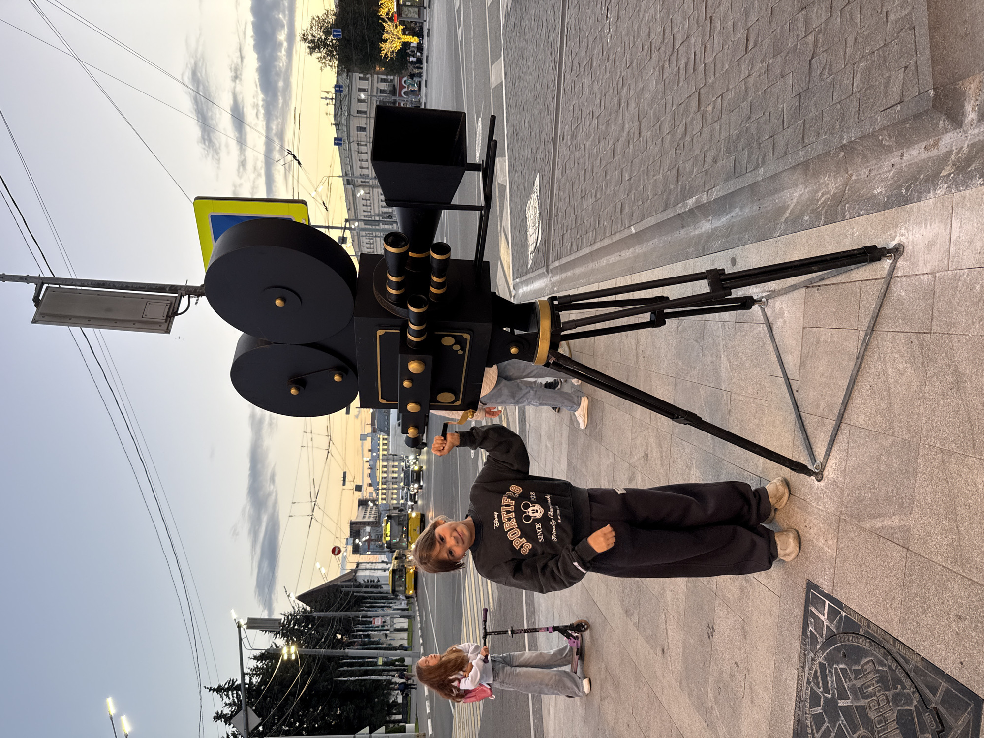 A street-level, eye-level medium shot captures a giant, black, vintage film camera prop on a tripod in a city square. The camera is positioned in the foreground, slightly off-center to the right. To the left of the camera, a young girl with long brown hair, wearing a black sweatshirt with "SPORTIFS" and a Mickey Mouse logo, and dark sweatpants, stands with her right arm raised, holding a black stick. She is smiling and looking towards the camera. Behind her, another young girl with long brown hair, wearing a white sweatshirt and light gray pants, stands with a pink backpack and holds onto the handlebars of a purple scooter. In the background, a street scene unfolds with traffic, including yellow buses and cars. Buildings with architectural details line the street, and overhead wires crisscross the sky. Streetlights and a decorated tree with lights are visible in the distance. The ground is paved with gray and beige square tiles. The sky is a pale blue with wispy clouds, indicating dusk. The location is the city of Zhukovo, Russia.