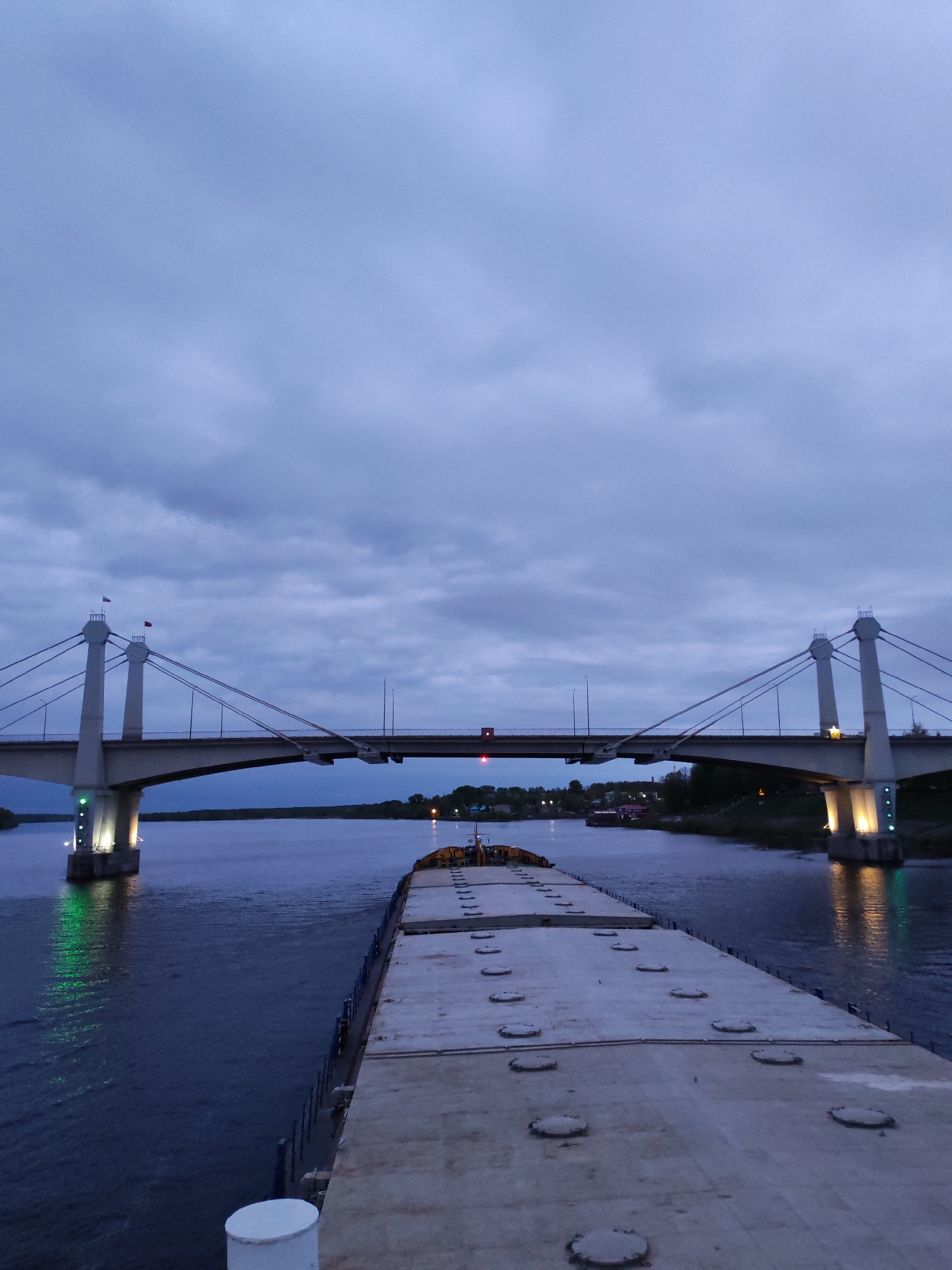 The image depicts a view from the deck of a barge navigating a river toward a bridge in the city of Kimry, Russia. The sky is overcast with a variety of cloud types. The bridge is a cable-stayed structure with two prominent towers. Lights illuminate the bridge supports and reflect in the water. A second bridge span is visible behind the primary one. The barge is long and flat with a repeating pattern of access covers. Vegetation lines both banks of the river. A few buildings and lights are visible in the distance. The overall scene is captured at dusk or dawn.