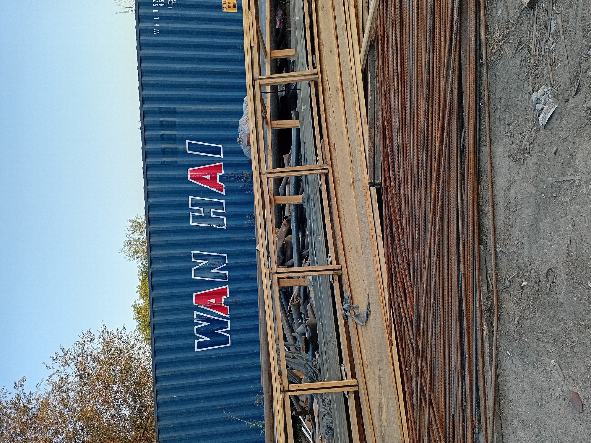 The image depicts an outdoor scene with a large blue shipping container prominently displayed in the background, bearing the white and red lettering "WAN HAI". In the foreground, a jumble of construction materials is visible, including stacks of wooden planks and a significant quantity of rusted rebar. A temporary wooden structure, possibly a makeshift support or railing, is positioned in front of the container, partially obscuring some of the materials behind it. The ground surface appears to be a mixture of dirt and debris. The overall impression is of a construction site or a storage area for building supplies. The sky is clear and blue, suggesting daylight hours. The location in the city of Semey, Kazakhstan is noted.