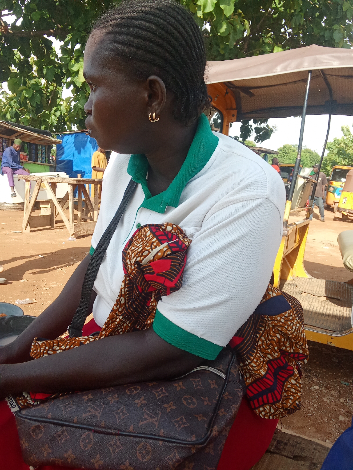 A medium-shot profile of a Black woman with braided hair is captured outdoors in a market setting. She is wearing a white polo shirt with a green collar and trim, and a colorful patterned fabric is draped over her shoulder. A dark-colored handbag with a monogram pattern rests on her lap, and a dark strap is visible across her chest. In the background, a yellow and brown motorized tricycle, market stalls, and other individuals are present. The ground is dirt, and there is foliage from trees above. The scene appears to be in an urban environment.