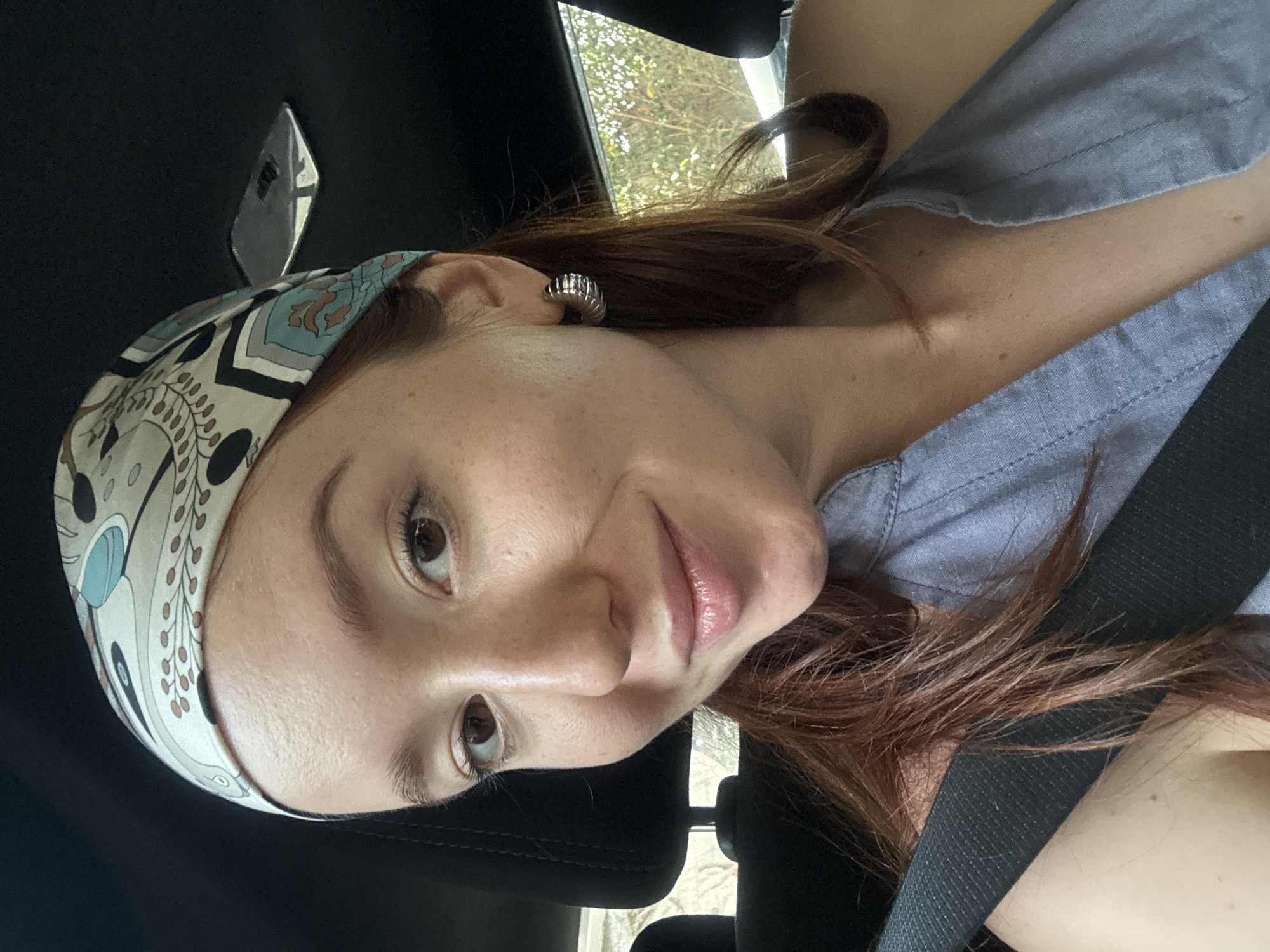 A person with reddish-brown hair, wearing a patterned headscarf and a light grey shirt, is sitting in a car. They are wearing a seatbelt and an earring. The location is inside a vehicle, not identifiable as Choeng Thale, Thailand, based on the image alone.