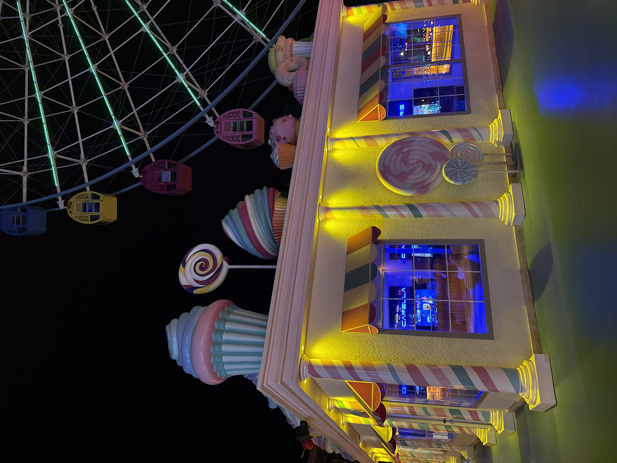 The image depicts a whimsical, candy-themed amusement park or attraction at night. A large Ferris wheel, illuminated with green lights, dominates the upper right portion of the frame. Below the Ferris wheel, a brightly colored building resembling a candy store or cafe is visible. The building's facade is painted in pastel yellow and decorated with striped columns and awnings. Oversized candy decorations, such as cupcakes and lollipops, are displayed on the roofline and in front of the building. The windows of the building are illuminated with blue light, revealing interior decorations or displays. The ground in front of the building is a dark, reflective surface, possibly a pathway or plaza, with reflections of the building's lights. The overall atmosphere is festive and playful, likely a commercial entertainment venue. The scene is set in Bodrum, Turkey, as indicated by the caption. There are no visible people in the image.