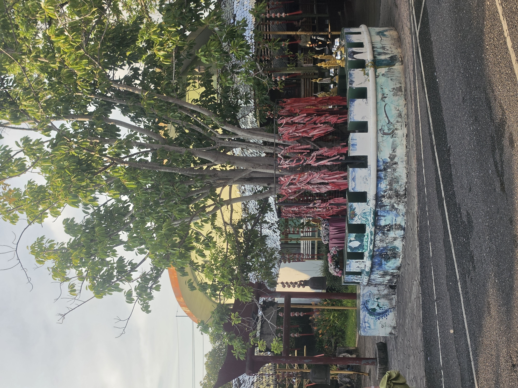 An outdoor scene in Pattaya City, Thailand, is depicted, showing a blend of natural and man-made elements. The image, originally rotated, features a dark asphalt road with two white parallel lines on the right side. Adjacent to the road is a low, curved wall or barrier, richly decorated with intricate blue, white, and green painted patterns that may depict aquatic life or mythical figures. Along this wall, numerous red strips of fabric or paper, resembling prayer ribbons or wish tokens, are tied, adding a splash of color.
A large, leafy tree with dense green foliage and thick branches dominates the left and center of the frame, providing shade. Behind the wall and amidst the tree's branches, glimpses of blue water are visible, suggesting proximity to a sea or large river. Various cultural and decorative objects are scattered in the midground, including small golden statues, rows of hanging bells or gongs, and other traditional ornaments, indicating a temple, shrine, or culturally significant site. A large, curved structure, colored in yellow and orange hues, is partially visible in the background, possibly a decorative archway or part of a building.
The bright sky, seen through the tree canopy, suggests it is daytime, possibly morning or late afternoon, with soft, diffused light. The scene appears peaceful and culturally rich, devoid of any visible people. No distinct, legible text is apparent on any of the objects.
