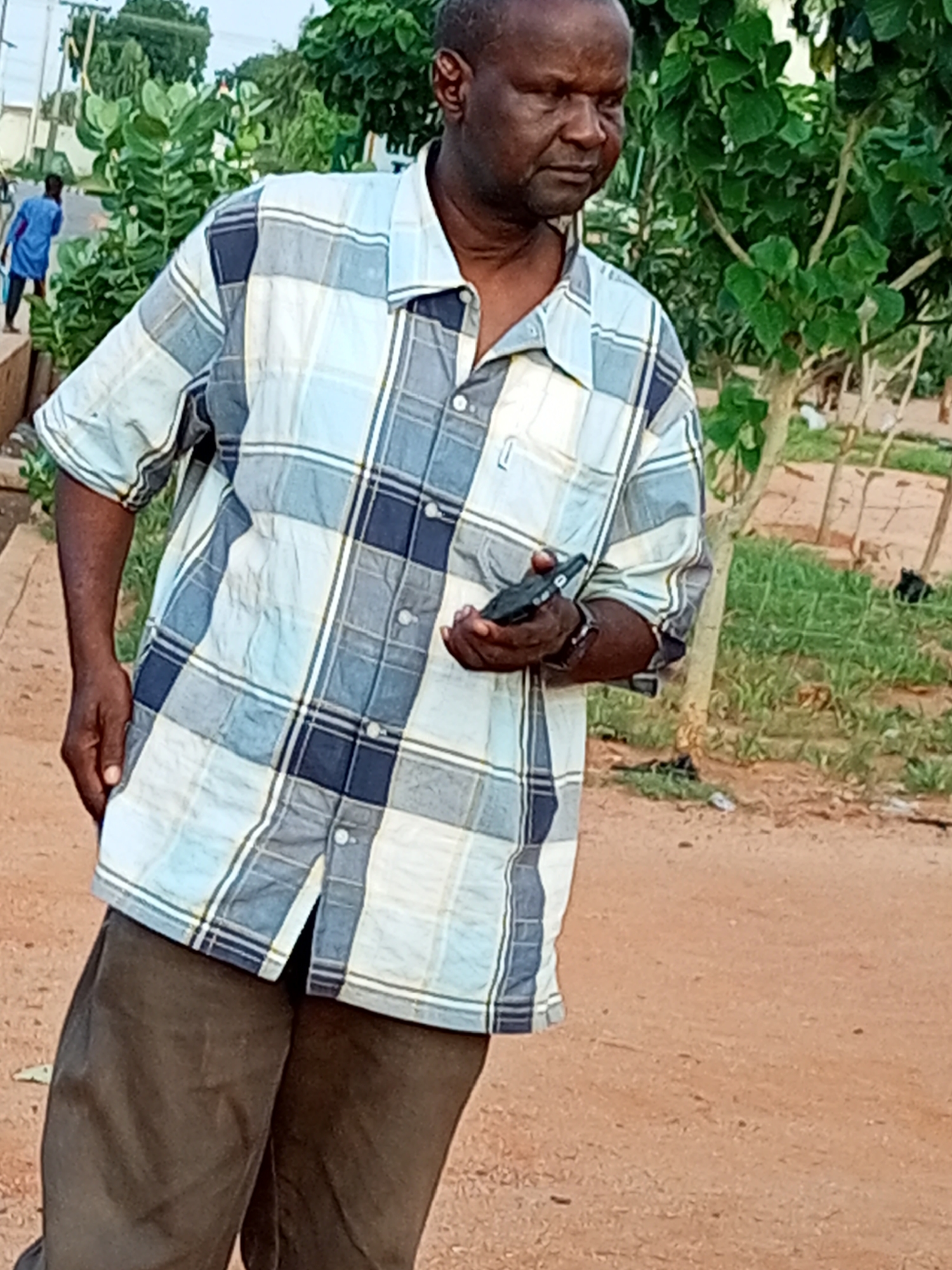 The image captures a medium shot of an adult Black male standing outdoors in a casual setting. He is wearing a plaid short-sleeved button-down shirt in shades of blue, white, and dark blue, with brown pants. The man is holding a black flip phone in his right hand, with his left arm bent and resting near his side. His gaze is directed slightly to his right, with a neutral expression.
The background suggests an outdoor urban or semi-urban environment. To the left, a person wearing a blue shirt and black pants is visible walking away from the camera. Lush green foliage from trees and bushes is prominent throughout the background. The ground appears to be a mixture of reddish-brown dirt and some patches of dry grass. The overall lighting indicates it is daytime.
The image is likely from Gombe, Nigeria, based on the visual cues of vegetation, ground cover, and the style of clothing worn by the individuals, which are consistent with common observations in the region. The scene depicts a casual moment, possibly a street or park area.