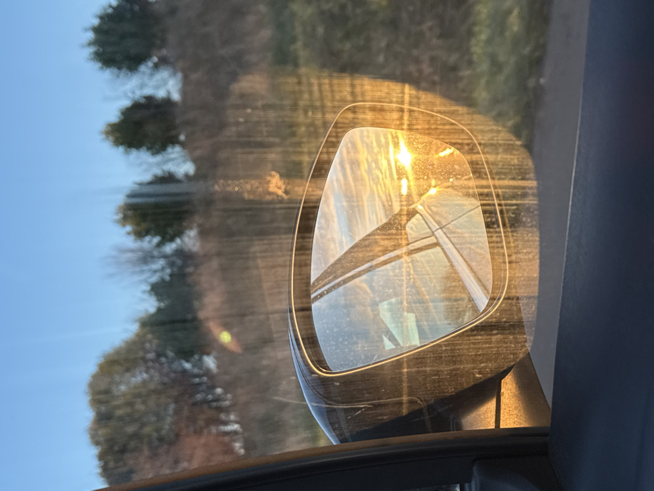A car's side mirror reflects sunlight and what appears to be power lines or tall structures against a bright sky. The mirror's surface is dusty or dirty, obscuring the reflection slightly. The background outside the car window shows trees and blue sky, adding depth to the composition. The image is taken from inside the car, showing part of the door frame.