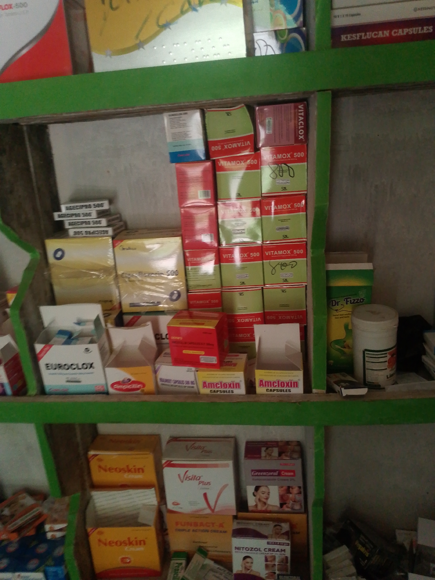 This media file captures a close-up, slightly angled view of a shelf filled with various pharmaceutical products in a store in Jauro Yinu, Nigeria. The shelves are painted green, and the products are primarily displayed in cardboard boxes and plastic containers. The items include medications like "VITAMOX 500," "VITACLOX," "EUROCLOX," "Amcloxin," "Neoskin," "Visita Plus," and "FUNBACT-A." There are also containers with labels such as "Dr. Fizzo" and "Greenzoral Cream." Some boxes are stacked neatly, while others are partially opened or arranged in a less orderly fashion. The lighting suggests it might be daytime, possibly indoors with artificial lighting contributing to the overall illumination. The scene appears to be a retail environment, likely a pharmacy or a small shop selling medicines. Visible text on the packaging provides brand names, dosages, and product types. There are no people visible in the frame. The overall impression is a well-stocked but somewhat cluttered display of health and personal care products.