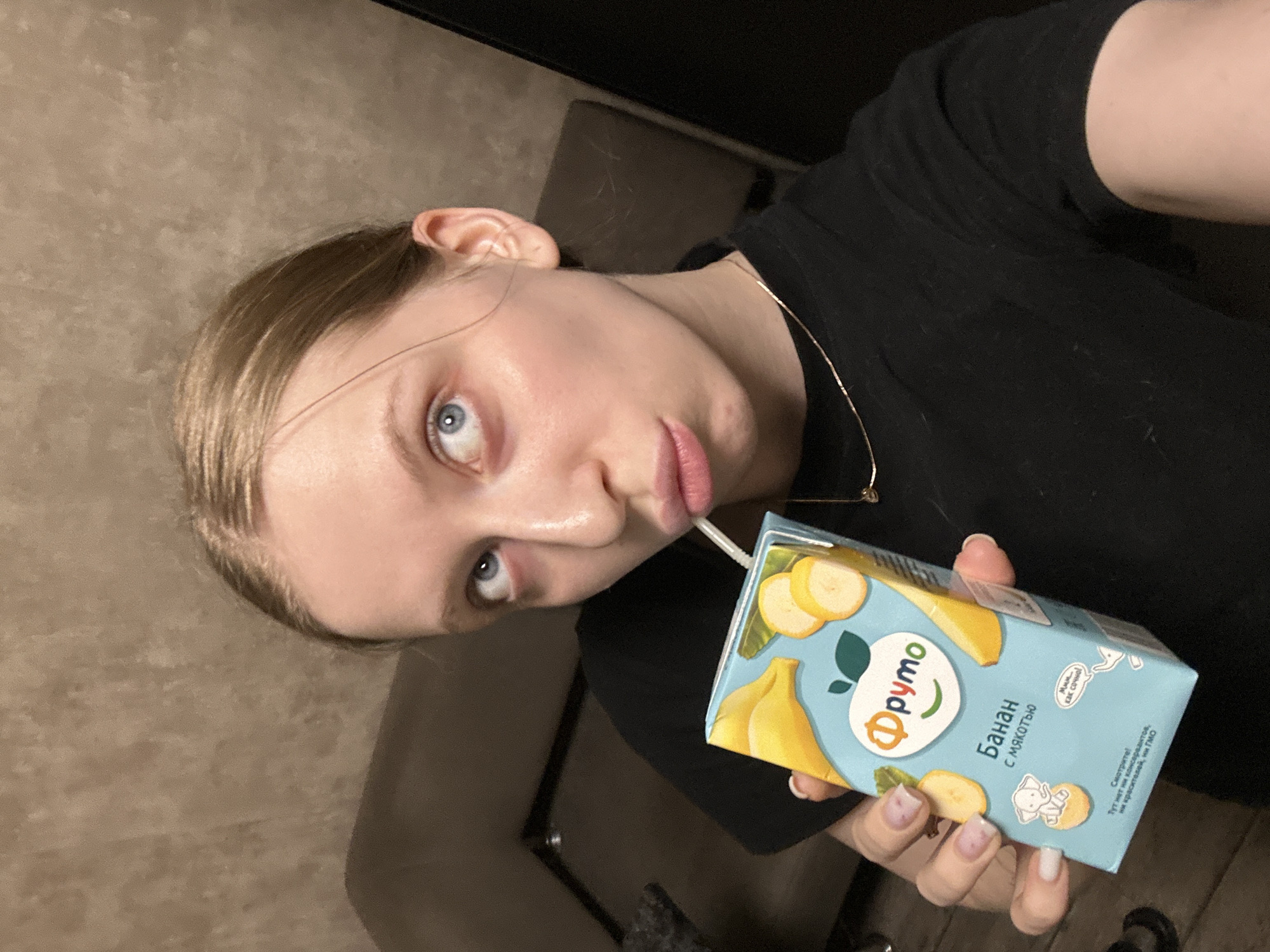 A young woman is drinking from a box of Fruto brand banana juice with a straw. The juice box is blue and features images of bananas, apples, and a cartoon elephant. The woman has blonde hair and blue eyes. She is wearing a black shirt and a gold necklace. The background is blurry, but it appears to be an indoor setting. There is a couch and a wall visible in the background. The juice box contains Russian text indicating "Banana with pulp." There is no explicit evidence to specify the location in Chaikovsky, Russia.