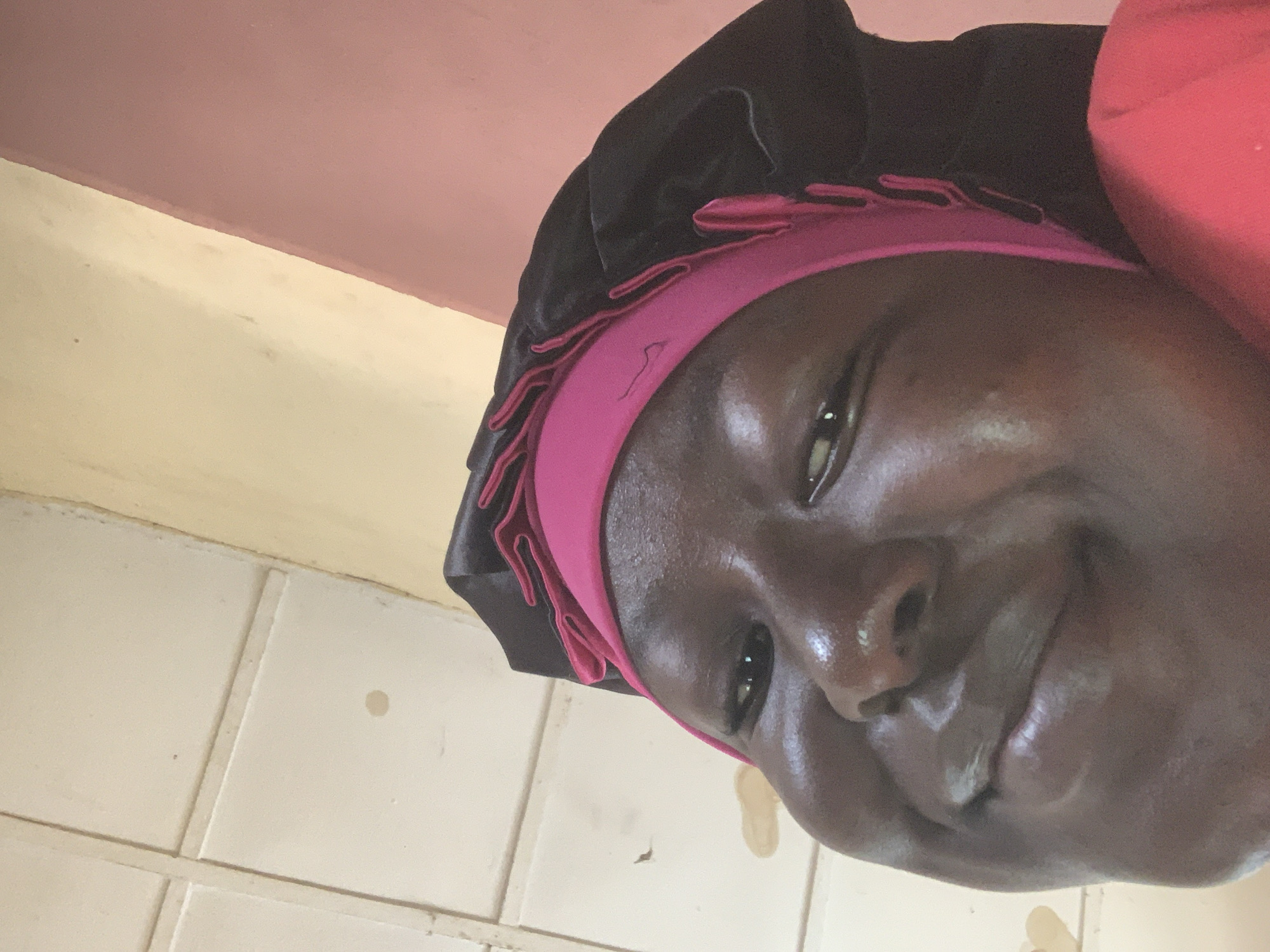 A low-angle, close-up selfie captures a young Black person with dark skin and a pink bonnet. The person is looking upwards at the camera with a slight smile. Their eyes are open, and their lips are slightly parted. The bonnet has a black satin exterior with a bright pink ruffled trim around the edge. The background consists of a textured ceiling with visible seams and a wall that transitions from a pale yellow to a muted pink. The lighting suggests an indoor environment, possibly a home. The specific location of Jauro Yinu, Nigeria is not identifiable from the visual elements in the image.