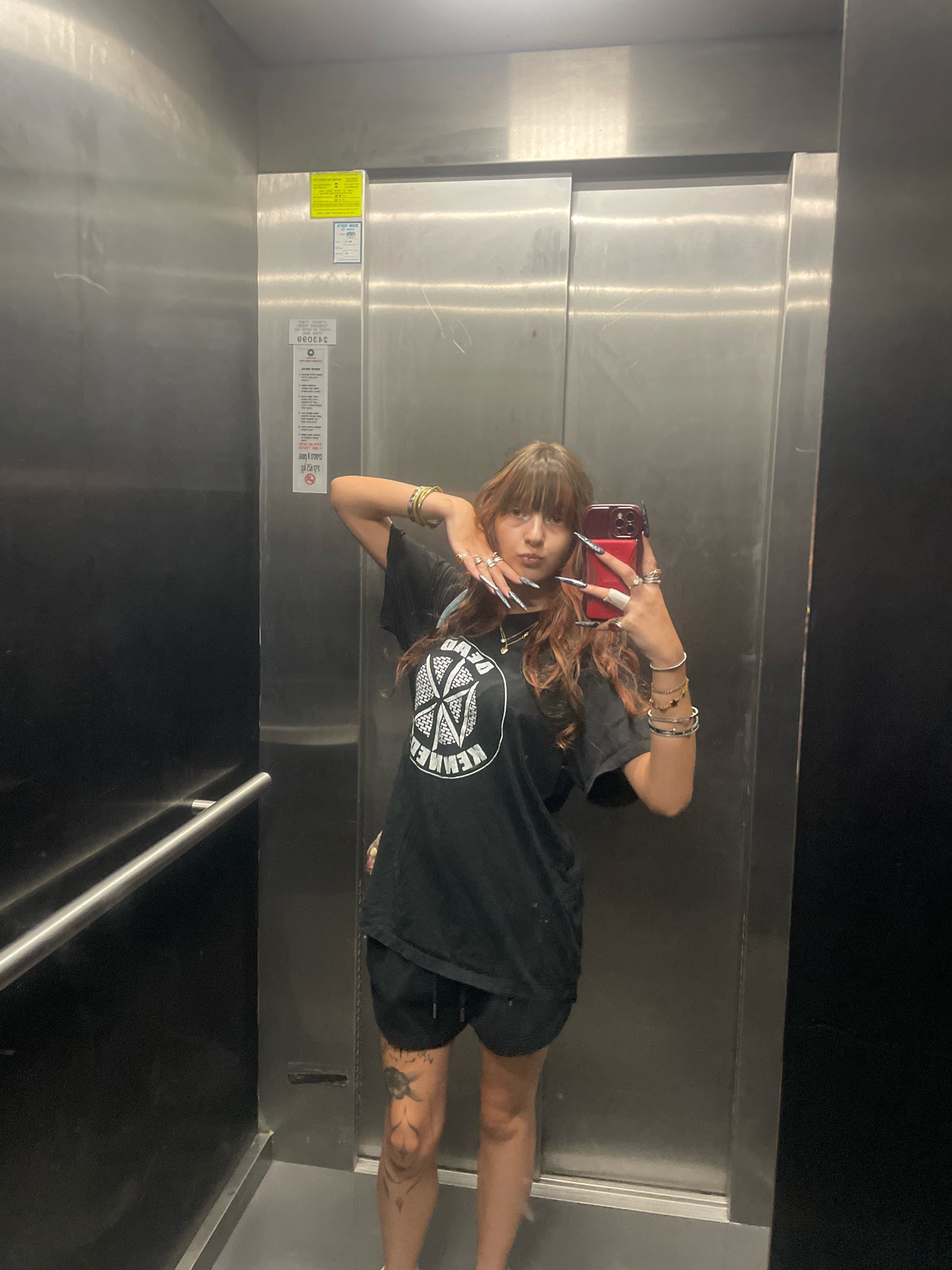 A young woman with long, dark, wavy hair and bangs takes a selfie in an elevator. She is wearing a black t-shirt with a white graphic on it and black shorts. She has long, sharp fingernails, multiple rings on her fingers, and several bracelets on her wrists. The elevator has stainless steel walls and doors, with a black panel on the left side containing a handrail. There are stickers on the elevator doors, including a yellow one at the top and a longer, white one below it. The floor of the elevator is a light grey. The overall environment appears to be a modern building. The sticker on the elevator door in the original image contains text that appears to be in Hebrew, and the presence of this script, along with the context provided, suggests the location is in Raanana, Israel.