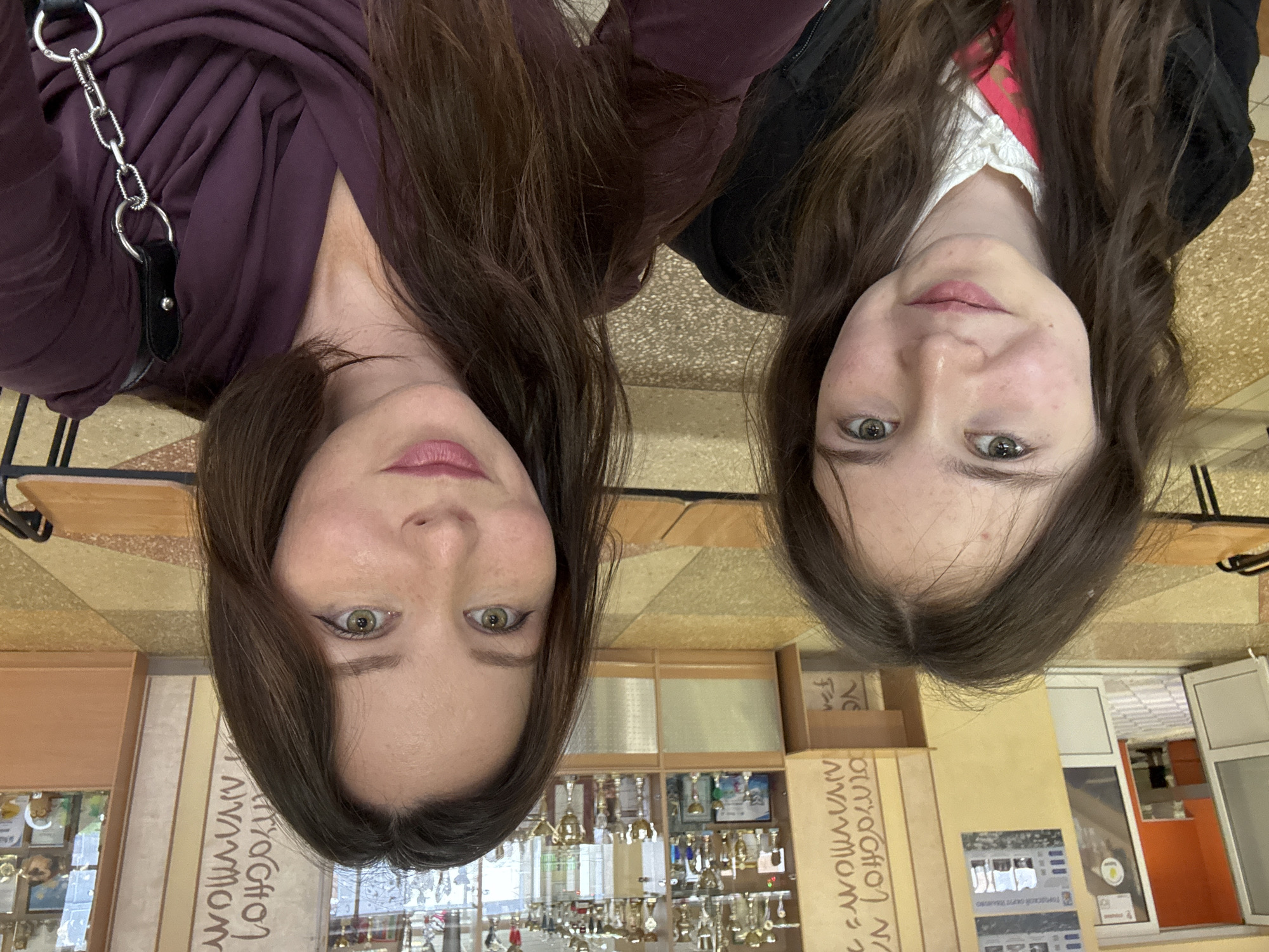 Two women are posing for a photo in what appears to be a school setting in Ivanovo, Russia. The image is rotated such that they are seemingly upside down.
On the left, a woman with dark hair is wearing a purple top and a bag with a silver chain. On the right, a younger woman with dark hair is wearing a white collared shirt with a pink top and a black jacket. Both women are looking directly at the camera.
The background includes shelves displaying various items and trophies, and a poster written in Russian. The floor has a tiled pattern. In the distance, one can see part of an orange wall and a window. There are also wooden tables and chairs visible.