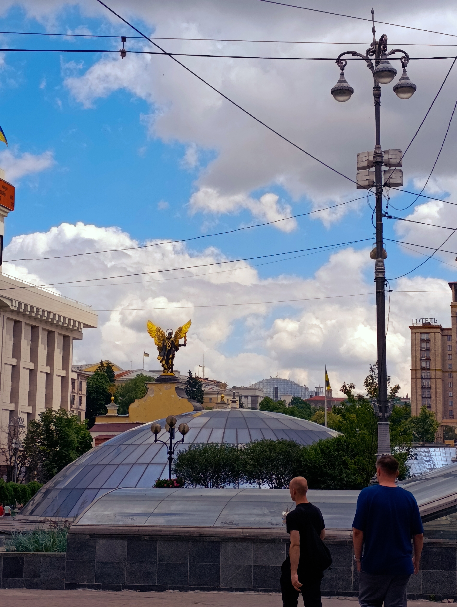 The image captures a wide shot of Maidan Nezalezhnosti (Independence Square) in Kyiv, Ukraine, under a bright, cloudy sky. In the foreground, two men walk away from the camera, one in a dark t-shirt and the other in a blue t-shirt and grey pants. Behind them, a large, domed glass structure, likely an entrance to an underground area, dominates the middle ground. Further back, the iconic golden statue of Archangel Michael, the patron saint of Kyiv, stands prominently on a pedestal, holding a sword and shield. To the right, a tall, multi-story building with the word "HOTEL" in Cyrillic script is visible. The scene is framed by overhead wires and a vintage-style lamppost. Green trees and other buildings are scattered throughout the background, indicating a bustling urban environment. The overall impression is one of civic space and historical landmarks.