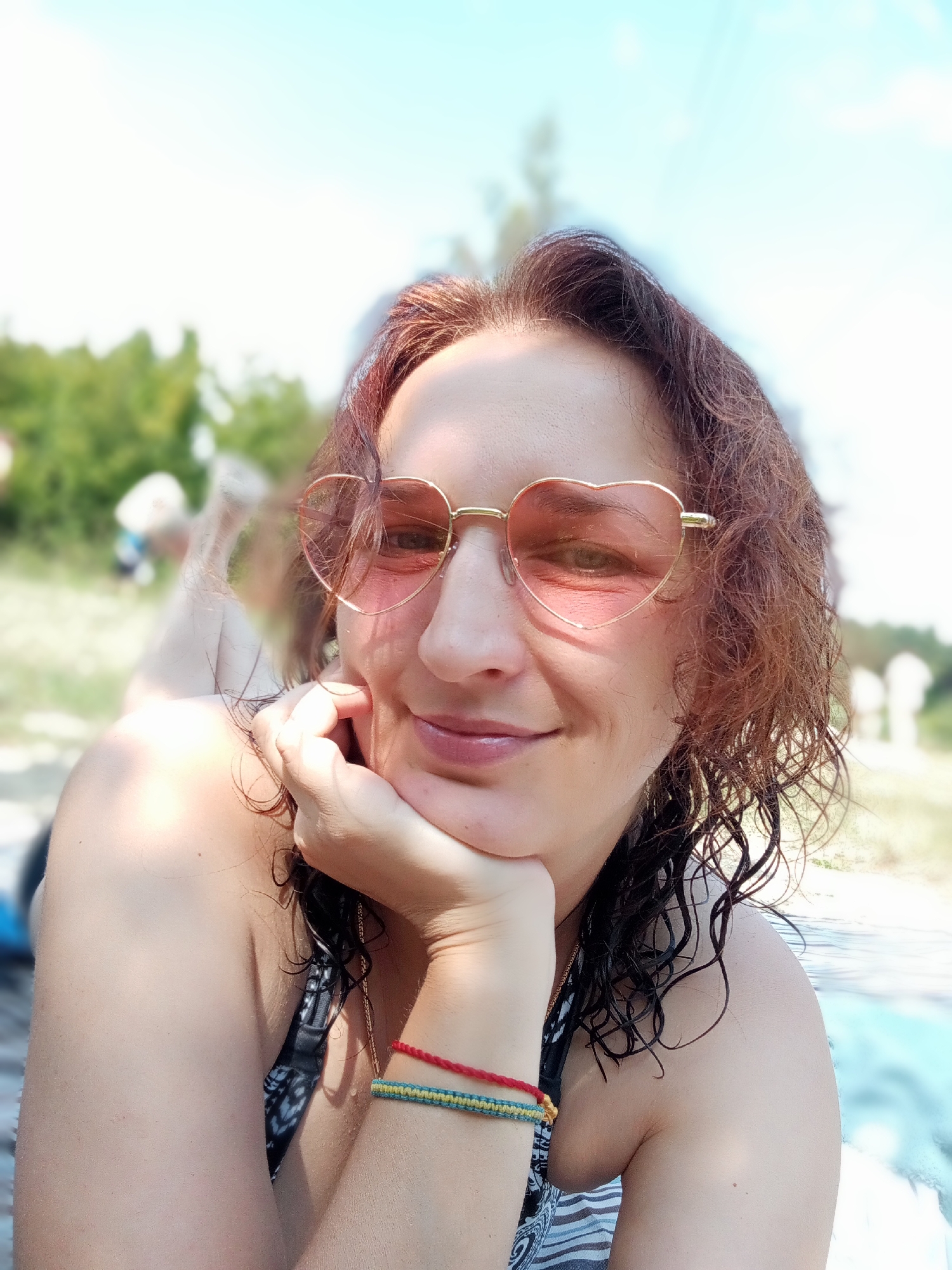 A woman with brown, wavy hair and heart-shaped pink sunglasses is lying down outdoors, propping her head up with her hand. She is wearing a black and white patterned bikini top. Two bracelets are visible on her left wrist: one red and one with blue and yellow threads. The background is blurred, showing trees and a body of water, suggesting a natural setting possibly near Boianivka, Ukraine. The lighting indicates a sunny day.