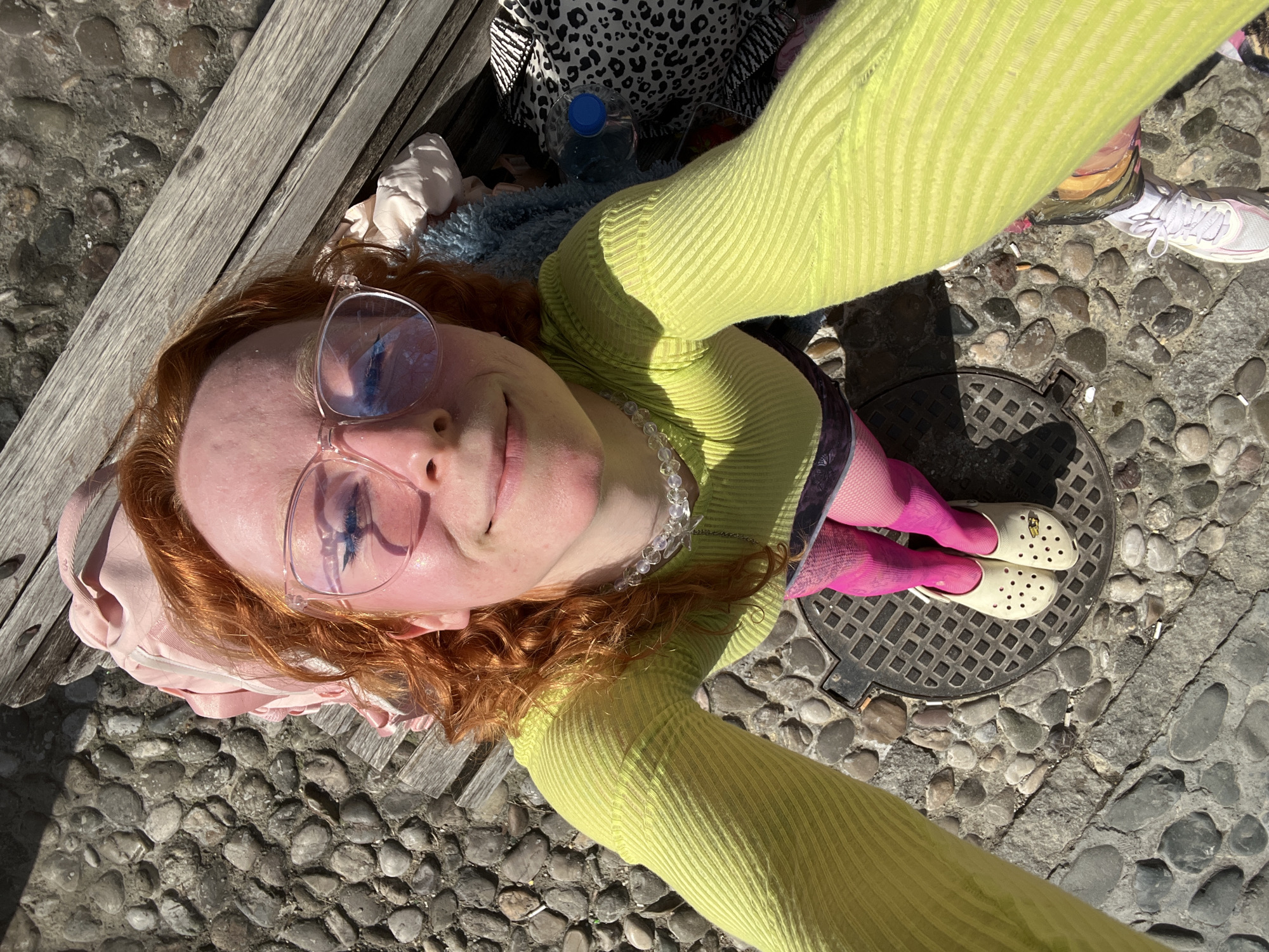 A selfie-style shot taken outdoors in what appears to be a pedestrian area in Belgrade, Serbia.
**Subject:** A young woman with reddish-brown curly hair and fair skin. She is wearing translucent frame glasses and a light green, long-sleeved ribbed shirt. She also has on pink tights or socks and white Crocs shoes with holes. She is positioned above a manhole cover.
**Objects:**
* Wooden bench (partially visible).
* Clothing items (likely a bag of clothes) next to the woman on the bench.
* Water bottle with a blue cap.
* Pearl or beaded necklace around her neck.
* Pebble stone ground surface.
* Manhole cover with a square grid pattern.
* A white sneaker or running shoe.
* Cigarette butts scattered on the ground.
**Environment:**
* The photo is taken in daylight, with shadows suggesting sunny conditions.
* The background appears to be a pedestrian street or square in an urban setting, possibly in Belgrade, Serbia, based on the architecture and cobblestone ground.