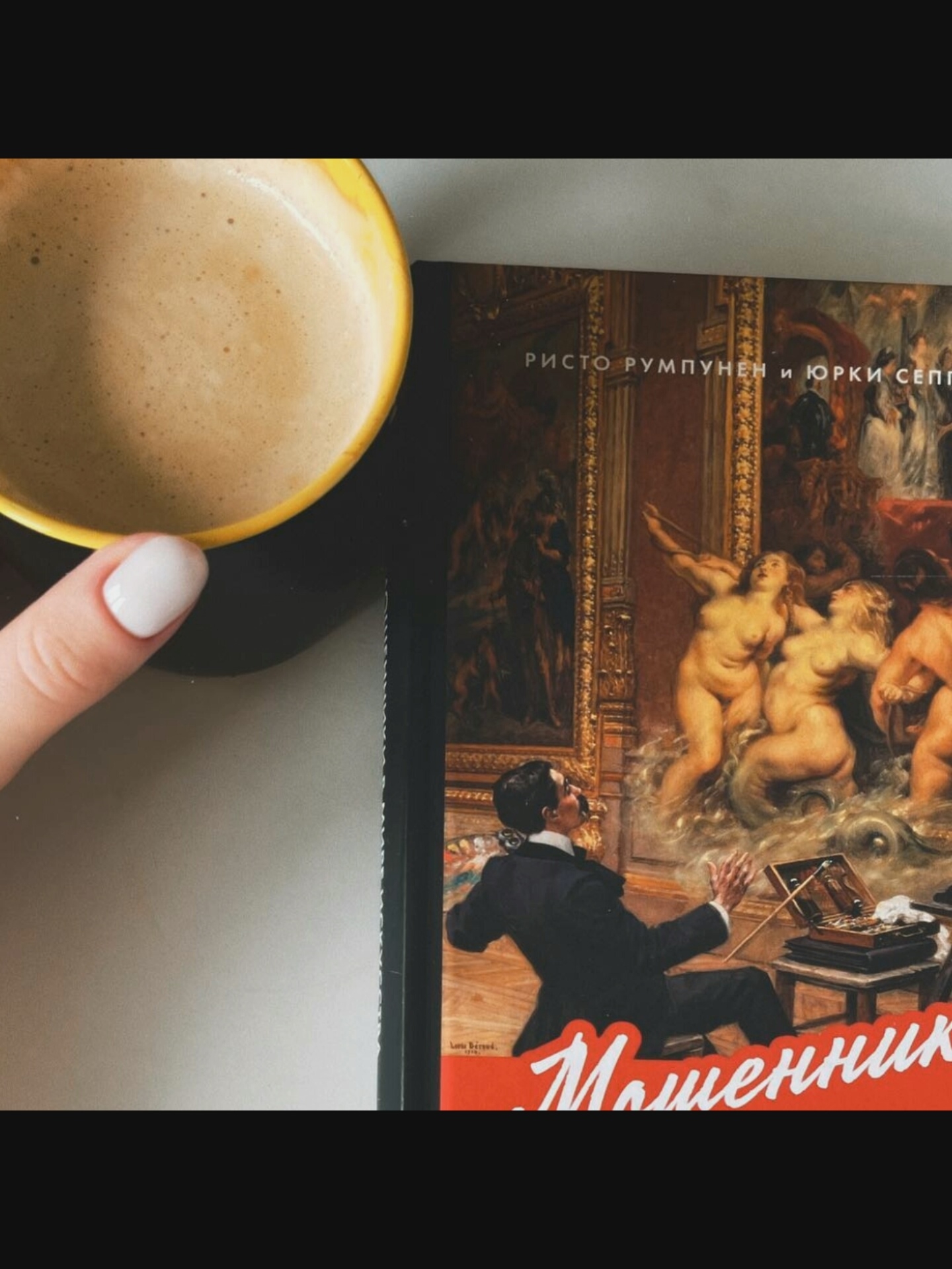 A top-down, slightly angled shot shows a yellow mug filled with a light brown, frothy beverage positioned on the left side of the frame. A person's left hand, with neatly manicured white fingernails, is visible at the bottom left, touching the rim of the mug. To the right of the mug is a book with a painting on its cover. The painting depicts several nude figures in a swirling, watery environment, with a man in a suit in the foreground looking upwards with his hands raised. Text in a stylized white font is overlaid on the bottom of the book cover. The background appears to be a dark surface, possibly a table, with a thin vertical black line running along the edge. The overall scene is dimly lit. There is no indication of the city of Saint-Août, France, or any specific location.