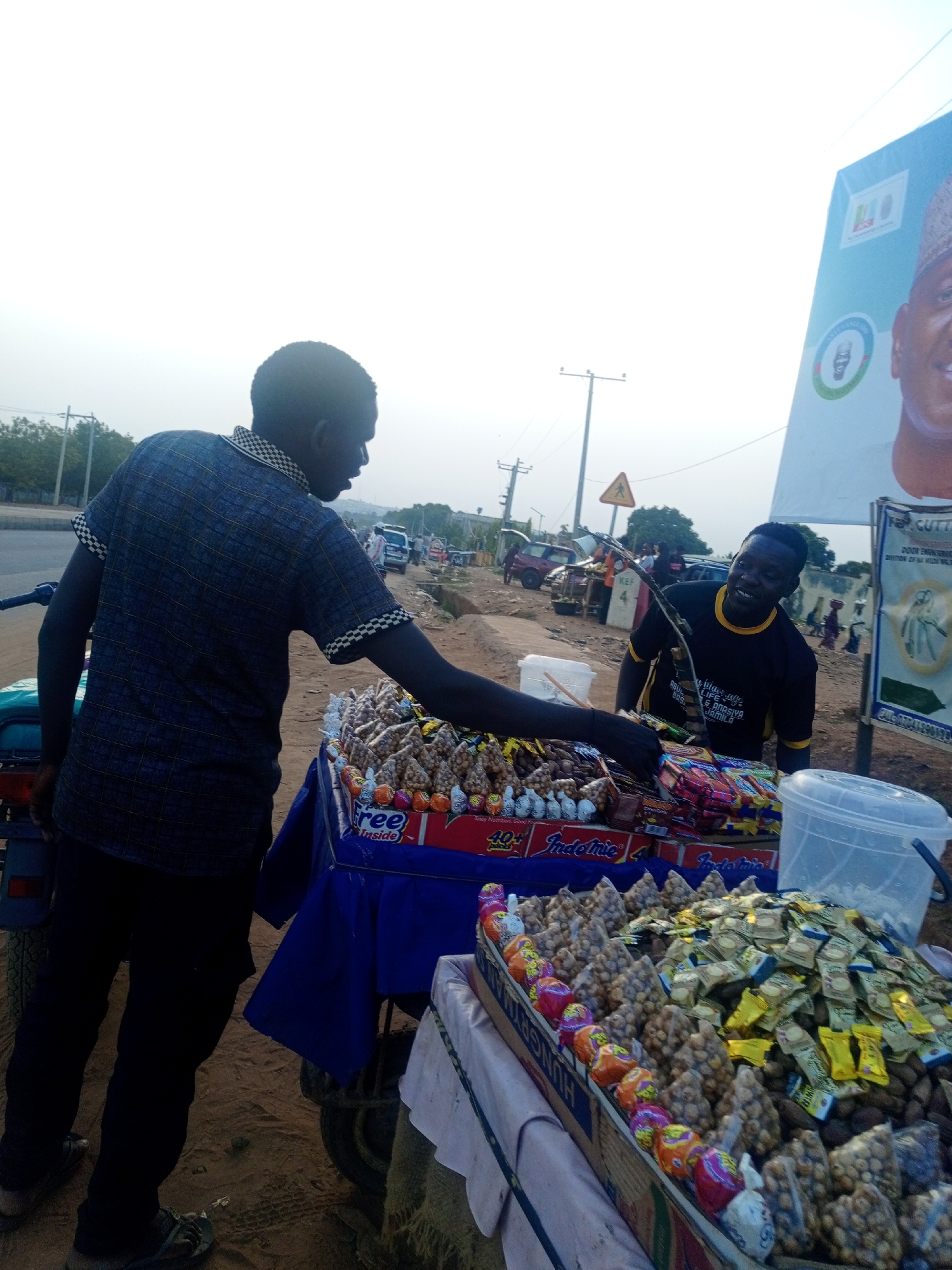 Two men are engaged in an interaction at a roadside snack stall, likely in Keffi, Nigeria, given the contextual hint. The man on the left, viewed from behind, wears a dark blue checkered shirt and dark trousers, reaching for items displayed on the stall. The vendor, on the right, smiles brightly at the camera, wearing a black and yellow trimmed t-shirt with some faded text.
The stall consists of two levels, heavily laden with various packaged snacks. Prominently displayed are numerous small, clear plastic bags containing nuts (possibly groundnuts or tiger nuts), arranged in pyramidal stacks. Colorful candies and other packaged goods, including boxes of "Indomie" instant noodles, are also visible.
The scene takes place outdoors on a bright, hazy day. The ground is dirt, transitioning to an asphalt road in the background where cars and other vehicles can be seen. Power lines and utility poles line the road. A notable detail is a large billboard on the right, featuring the portrait of a man, likely a political figure, and various logos and text. Visible text on the billboard includes "KEF", "ABUL", "LIFE", and "& ANASIYA JAMILA". On the snack packaging, "Indomie", "40+", and "Free Inside" are discernible. A yellow triangular road sign with a pedestrian crossing symbol is also in the mid-ground. The overall setting is a bustling, informal street market or vending point.