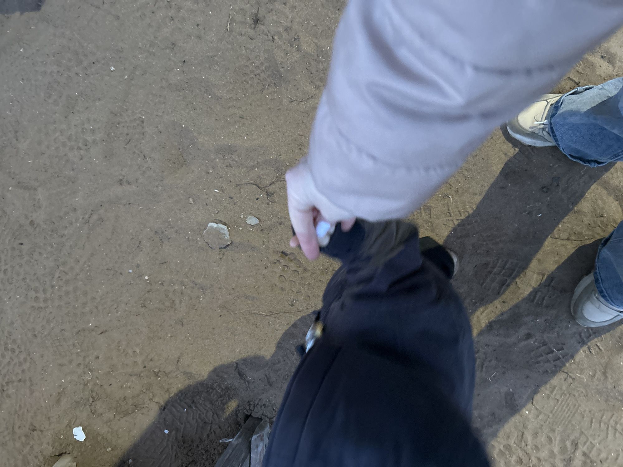 The image presents a top-down view of two individuals standing on a dirt surface in Noviy, Russia. The ground is uneven, marked with tire tracks or boot prints, and scattered with small pebbles and white debris. Strong directional lighting creates prominent, elongated shadows of the subjects.
On the left, the lower torso and left leg of one person are visible, clad in dark trousers. A small portion of a light-colored shoe is visible at the very bottom left. On the right, the lower torso and right leg of a second person are visible, wearing light blue jeans and light-colored sneakers. The right arm of this second person, covered by a light pink/beige coat sleeve, extends towards the first individual.
The right hand of the second person is grasping the hand or wrist area of the first person. At the point of interaction, a small metallic object, possibly a lighter, is visible against the dark fabric of the first person's clothing. The fingers of the second person's right hand appear to be holding a small, indistinct white object.