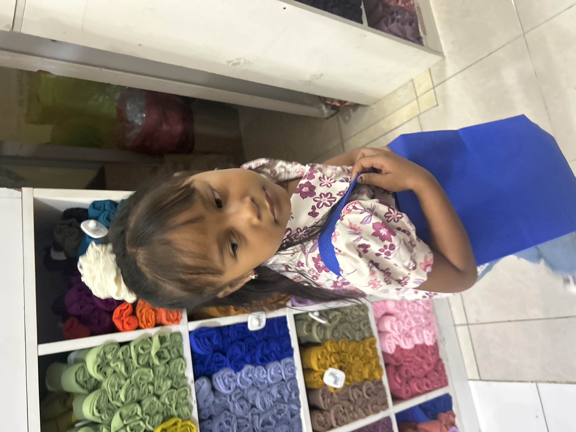 An eye-level, medium shot captures a young girl standing in front of shelves filled with rolls of fabric. The girl, with dark hair pulled back, wears a floral patterned shirt and holds a bright blue tote bag. She looks to her right with a neutral expression. The shelves behind her are stocked with a variety of colorful fabric rolls, ranging from light green and yellow to shades of blue, purple, and pink. To the right of the girl, a doorway or opening reveals more fabric rolls in red and green bags. The floor is tiled with light beige squares. The scene appears to be an indoor market or textile shop in Ubud District, Indonesia.