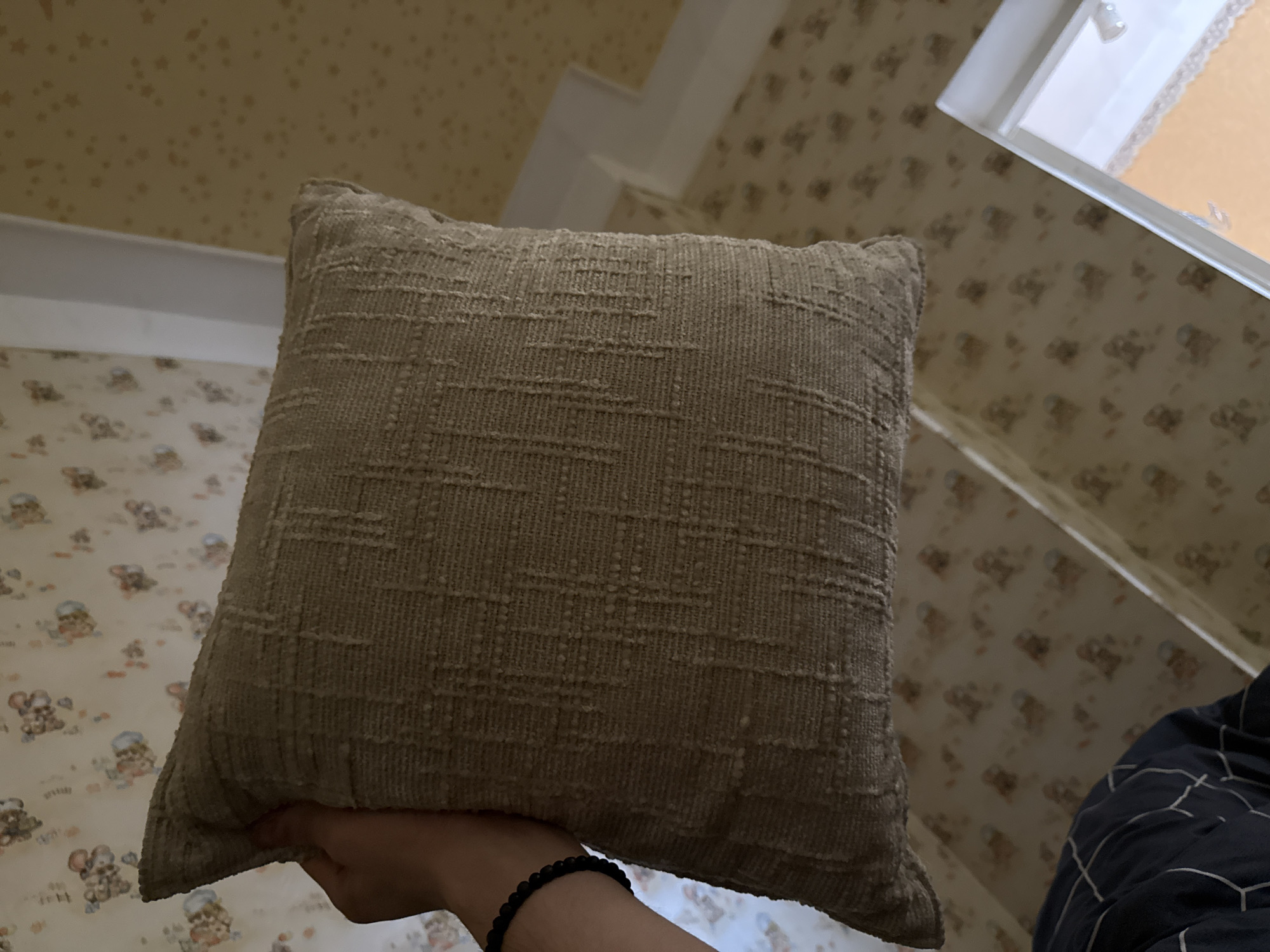 The media file shows a close-up, slightly angled view of a textured, tan throw pillow. The pillow is held by a person's hand, visible at the bottom left of the frame, and they are wearing a black beaded bracelet. The background features wallpaper with a repeating pattern of small, cartoonish bears wearing blue hats. Above the wallpaper, there is a white architectural element, possibly a ceiling molding or part of a doorway, against a textured yellow ceiling. A window frame is visible in the upper right, with what appears to be a light-colored curtain or blinds behind it. The lighting suggests it might be daytime, but the indoor setting offers no specific clues about the weather. The overall impression is of an indoor, domestic setting. No text is visible in the image.