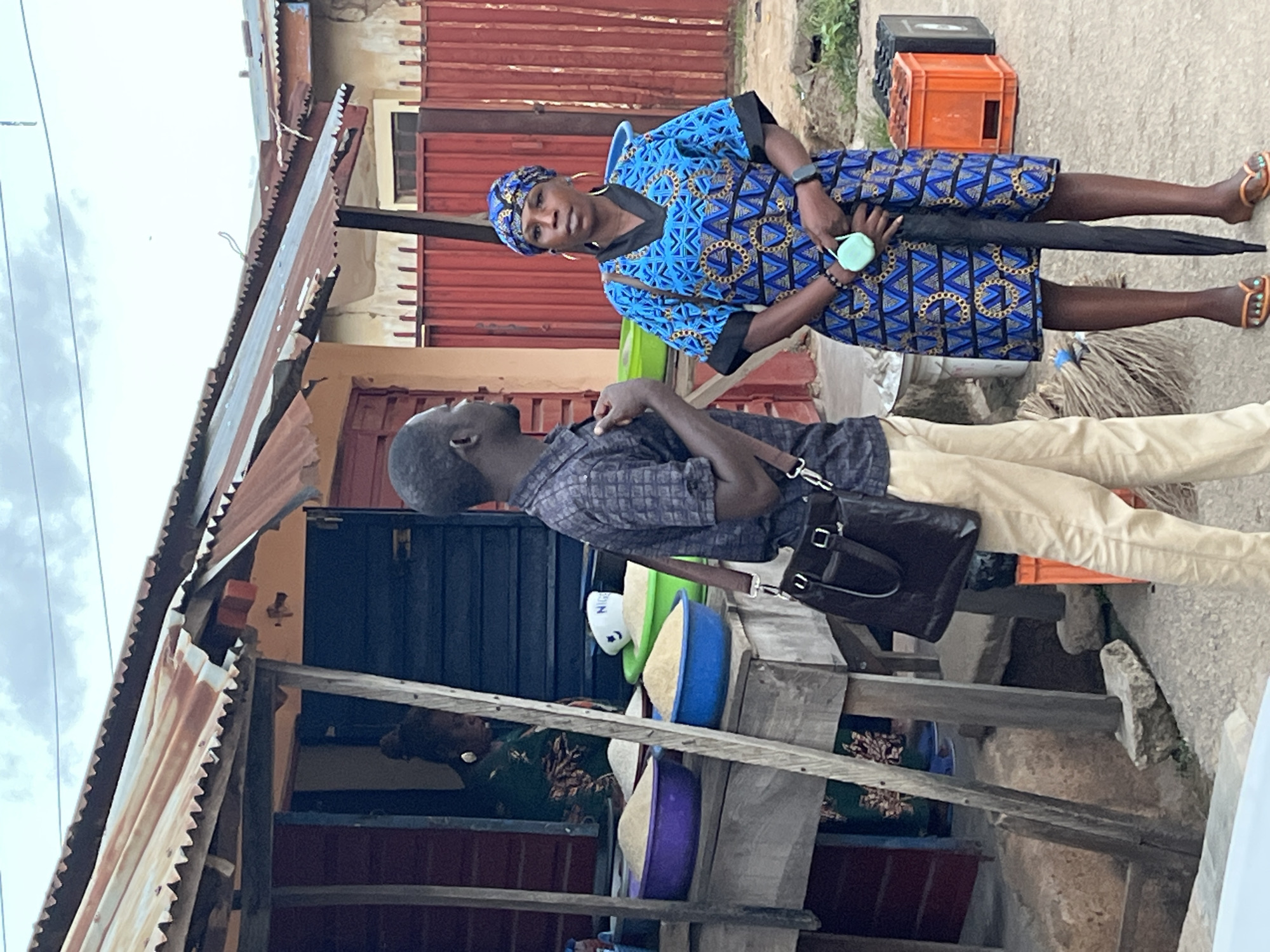 Two people are interacting outside a shop in Jos, Nigeria. One person is standing and dressed in a blue patterned dress, headwrap, and sandals. They are holding an umbrella and a small, round object. The second person is wearing a plaid shirt and khaki pants, and carrying a black bag. They are standing in front of a shop or stall, which has a tin roof, blue doors, and colorful bowls on a shelf. The environment suggests a commercial area with other buildings nearby.