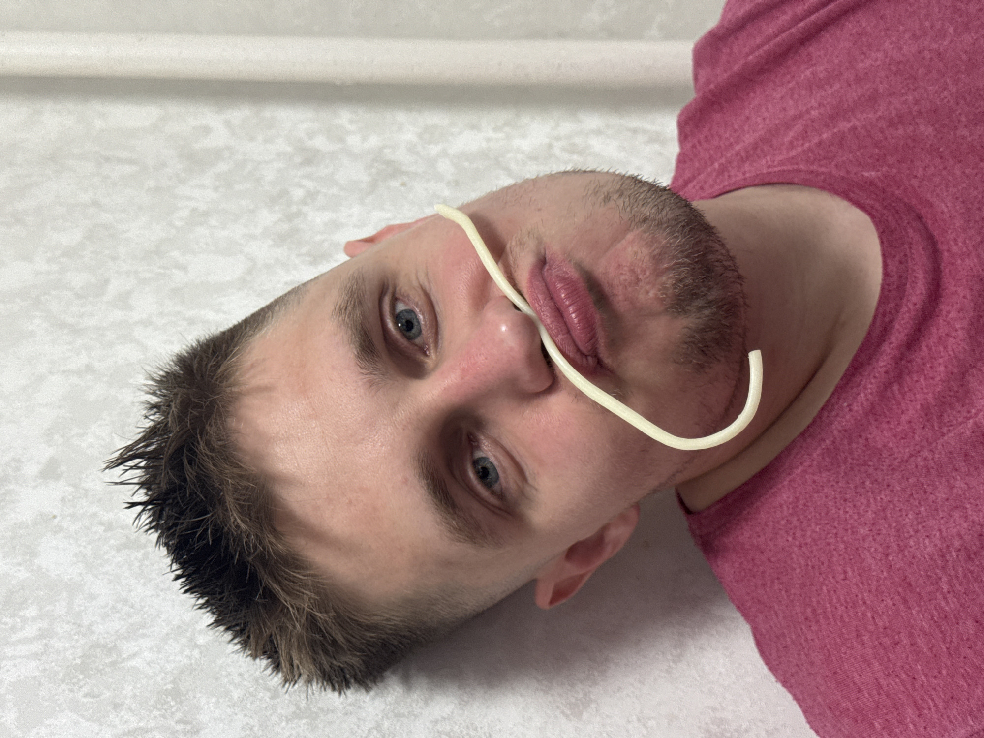 A close-up, head-and-shoulders shot shows a young man with short, spiky brown hair and blue eyes. He is wearing a bright pink t-shirt and leaning against a textured, off-white wall. A single strand of spaghetti is positioned across his upper lip, resting on his nose and curved downwards towards his chin, giving the appearance of a mustache. His lips are pursed. The lighting is direct, casting subtle shadows on his face. The background wall appears to be an interior surface, possibly a residential setting. The image does not provide any specific geographical or temporal indicators that would allow for its localization to Chaikovsky, Russia, or any other city.