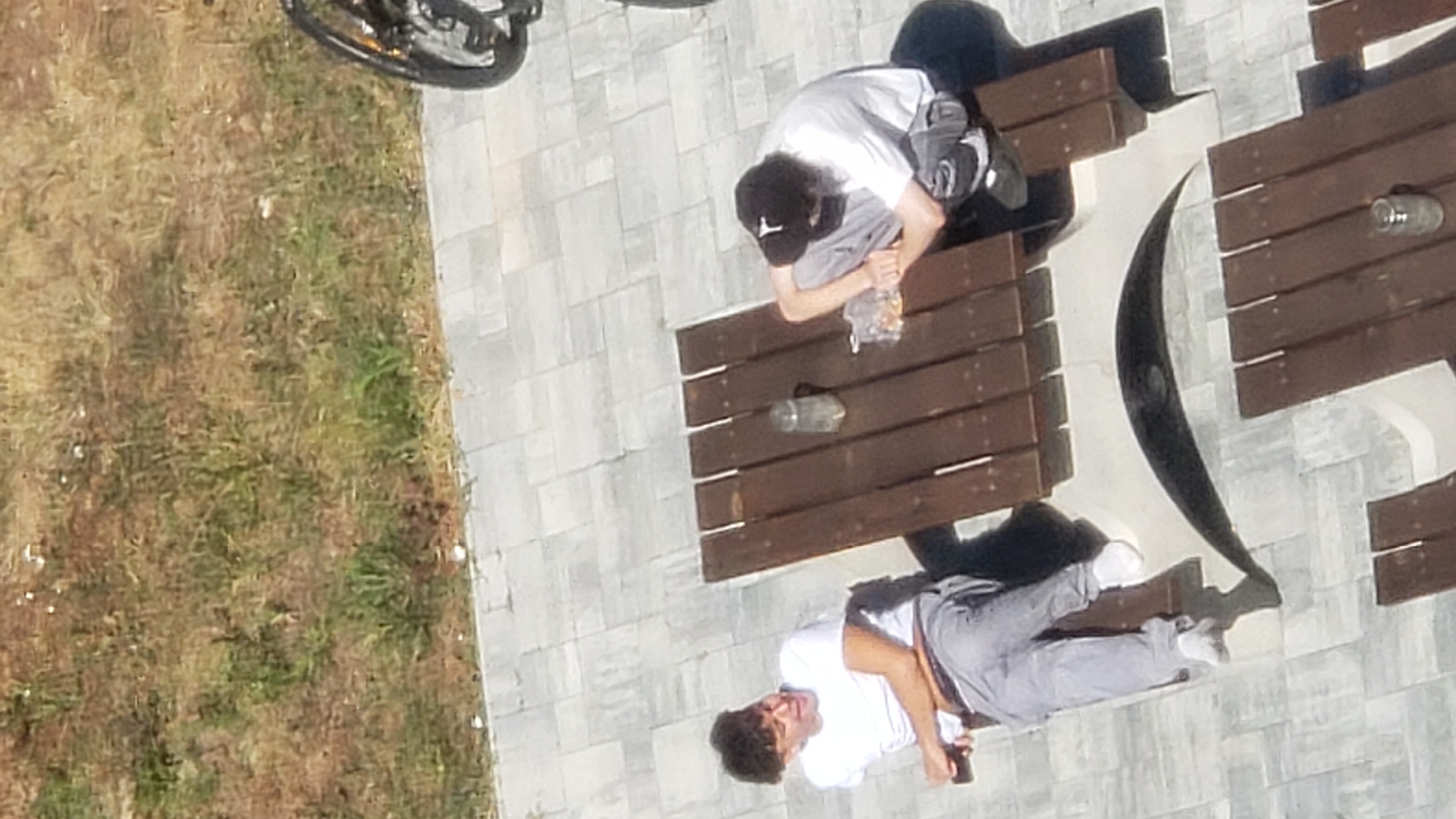 Two young men are seated at a wooden picnic table outdoors. The man on the left is wearing a white t-shirt and grey pants, and he is looking to his left. The man on the right is wearing a white t-shirt and grey pants, and he has a black baseball cap on. He is hunched over, looking at something in front of him. On the table between them, there is a glass jar and a plastic bag containing what appears to be snacks. The picnic table is situated on a paved patio with a patterned stone surface. In the background, there is a grassy area with scattered patches of dry grass and some green vegetation. To the right of the image, a portion of a bicycle wheel is visible. The overall scene suggests a casual outdoor setting, possibly a park or an outdoor seating area of a public establishment.