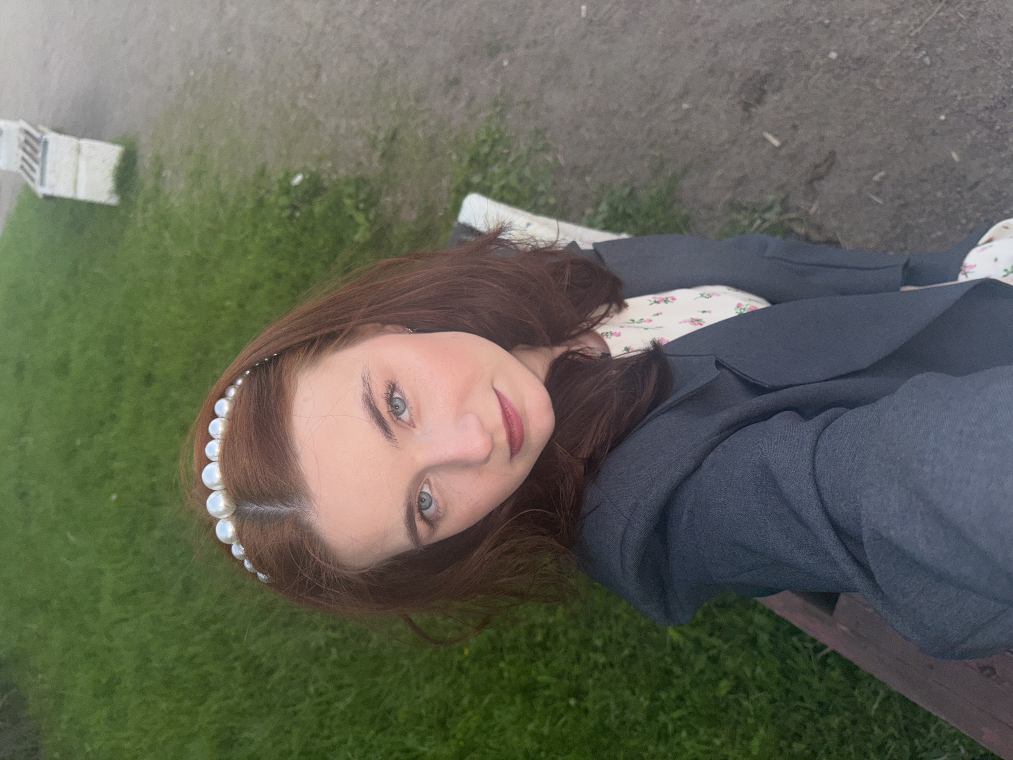 An overhead, slightly angled photograph captures a young woman from the chest up, seemingly reclining or leaning back on a wooden bench in an outdoor setting. She has long, reddish-brown hair styled with a pearl headband, and possesses fair skin, blue eyes, and lips colored with a muted red. Her gaze is directed upwards towards the camera, accompanied by a soft, pleasant expression and a slight smile.
She is dressed in a dark gray blazer or jacket worn over a light-colored top adorned with a delicate pink and green floral pattern. The wooden bench she occupies is visible beneath her and to her right. The background features a mix of green grass and a dirt or gravel path, characteristic of a park or garden. A faint white structure, possibly a fence or railing, can be seen in the distant upper left. The lighting is soft and diffused, suggesting an overcast day or indirect sunlight. The scene conveys a calm and serene mood. No text is visible in the image. The provided location context is Saint Petersburg, Russia.