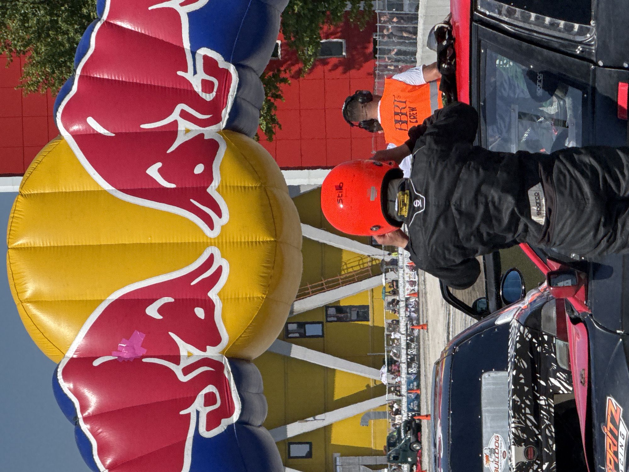 A close-up shot shows a person in a black racing suit and orange helmet leaning into a race car. Beside them is a crew member with an orange vest. In the background is a large inflatable Red Bull logo. The backdrop includes a building with yellow and red sections, and a crowd of spectators can be seen in the distance. The picture was taken during an auto racing event, possibly in the city of Plovdiv, Bulgaria.