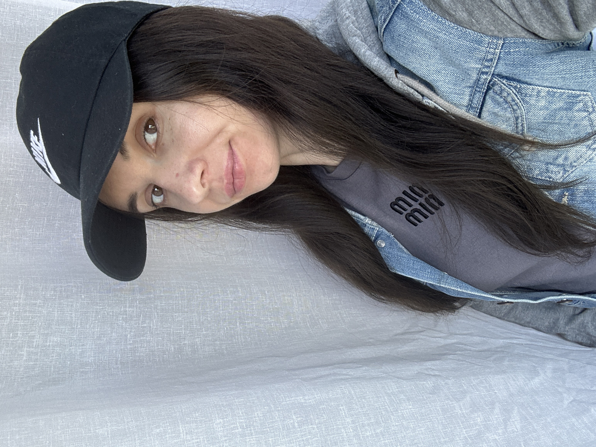 A woman with long, dark hair is wearing a black baseball cap with a white logo, a gray t-shirt with the word "MIUMIU" printed in black, a denim jacket, and a gray hoodie. She is tilting her head and smiling slightly at the camera. The background is a white textured surface. The location is unspecified.