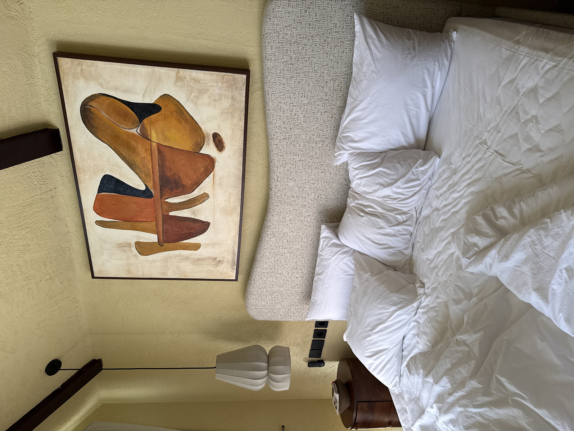 An overhead view captures an interior bedroom scene, likely in Kuta Selatan, Indonesia. The main focus is an unmade bed, occupying the right side of the frame, covered with white, rumpled sheets, a duvet, and several pillows. Behind the bed is a light-colored headboard featuring a distinct, repeating geometric pattern.
To the left of the bed, a dark wooden bedside table is visible, topped with a remote control and what appears to be a brown hat or cap. Above the table, a series of black rectangular electrical outlets or switches are mounted on the textured, warm yellow wall. A modern, multi-tiered white hanging lamp with a pleated design descends from the ceiling in the mid-left portion of the image.
On the far left, a framed abstract painting hangs on the wall. The artwork features large, organic shapes in shades of brown, ochre, and black, set against a lighter, textured background, framed by dark wood. A dark wooden beam runs diagonally across the upper left corner, possibly part of the ceiling structure. The ceiling, like the walls, is textured and painted in a similar warm yellow hue. No people are present. The scene suggests a relaxed and functional bedroom environment, possibly within a resort or hotel. There is no visible text in the image.