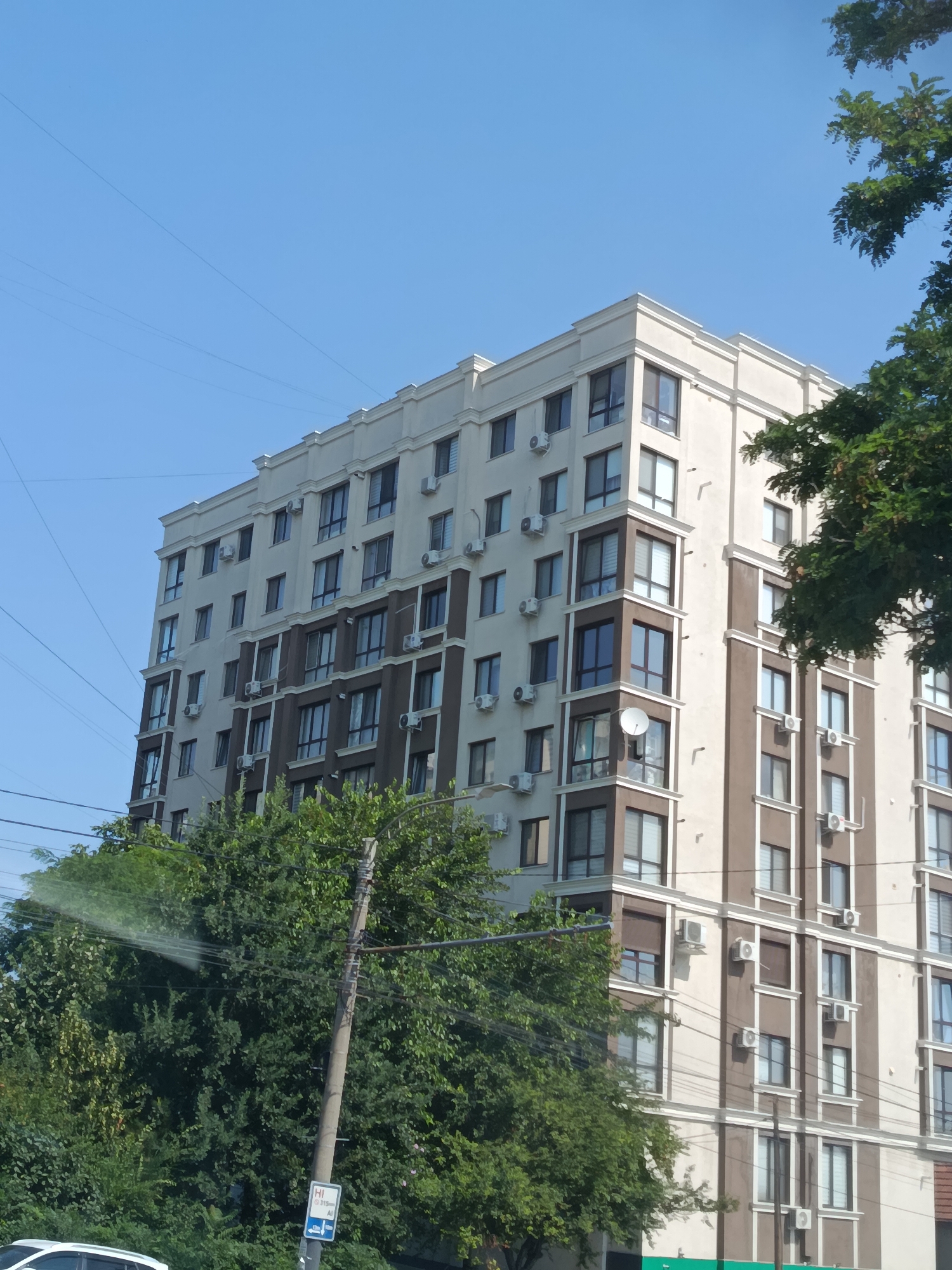 This is a low-angle, outdoor, daylight, eye-level, medium shot photograph of a modern, multi-story apartment building. The building has a light beige facade with dark brown vertical accents and white trim around the windows and balconies. The windows are large and rectangular, and many of them have white shutters or blinds. The building is covered in air conditioning units. Lush green trees surround the base of the building and partially obscure the lower floors. The sky is a clear, bright blue. Electrical wires are visible crossing the frame in the upper left and lower right quadrants. A utility pole with wires is prominent in the center of the lower portion of the image, with a small blue and white sign attached to it. A white car is partially visible in the bottom left corner. The scene appears to be in a residential area of Chișinău, Moldova.