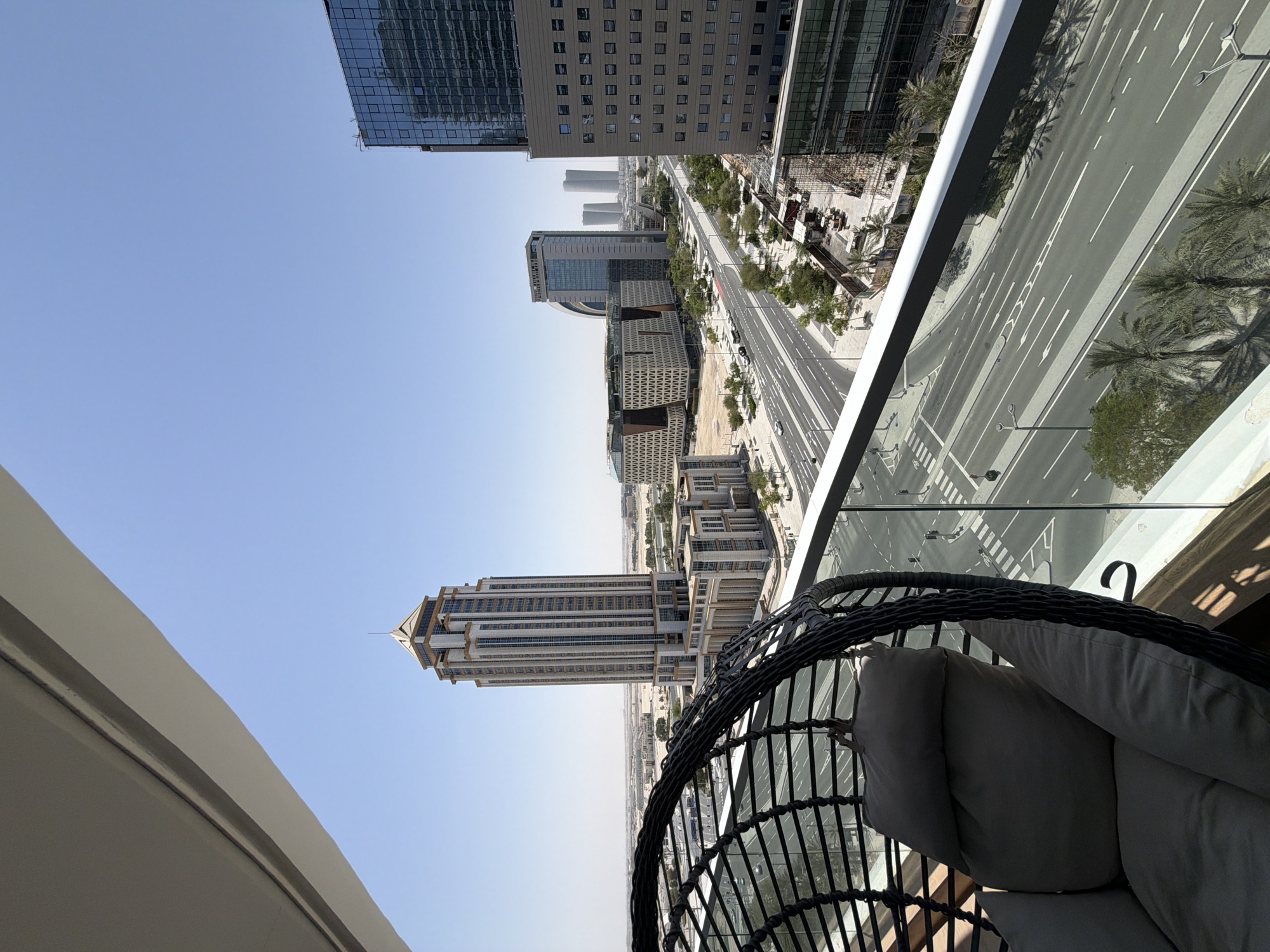This media file displays a high-angle view of a modern urban landscape. The scene is dominated by tall, contemporary buildings with glass facades and clean lines, interspersed with a wide, multi-lane road. Palm trees line the sides of the road, suggesting a warm climate. In the foreground, a dark, wicker hanging chair with light gray cushions is partially visible, implying a balcony or outdoor seating area. The sky is clear and a pale blue, indicating daytime with fair weather. The sunlight casts distinct shadows, suggesting it is likely mid-morning or early afternoon. The overall impression is one of a developed, possibly affluent urban environment. There is no visible text in the image.