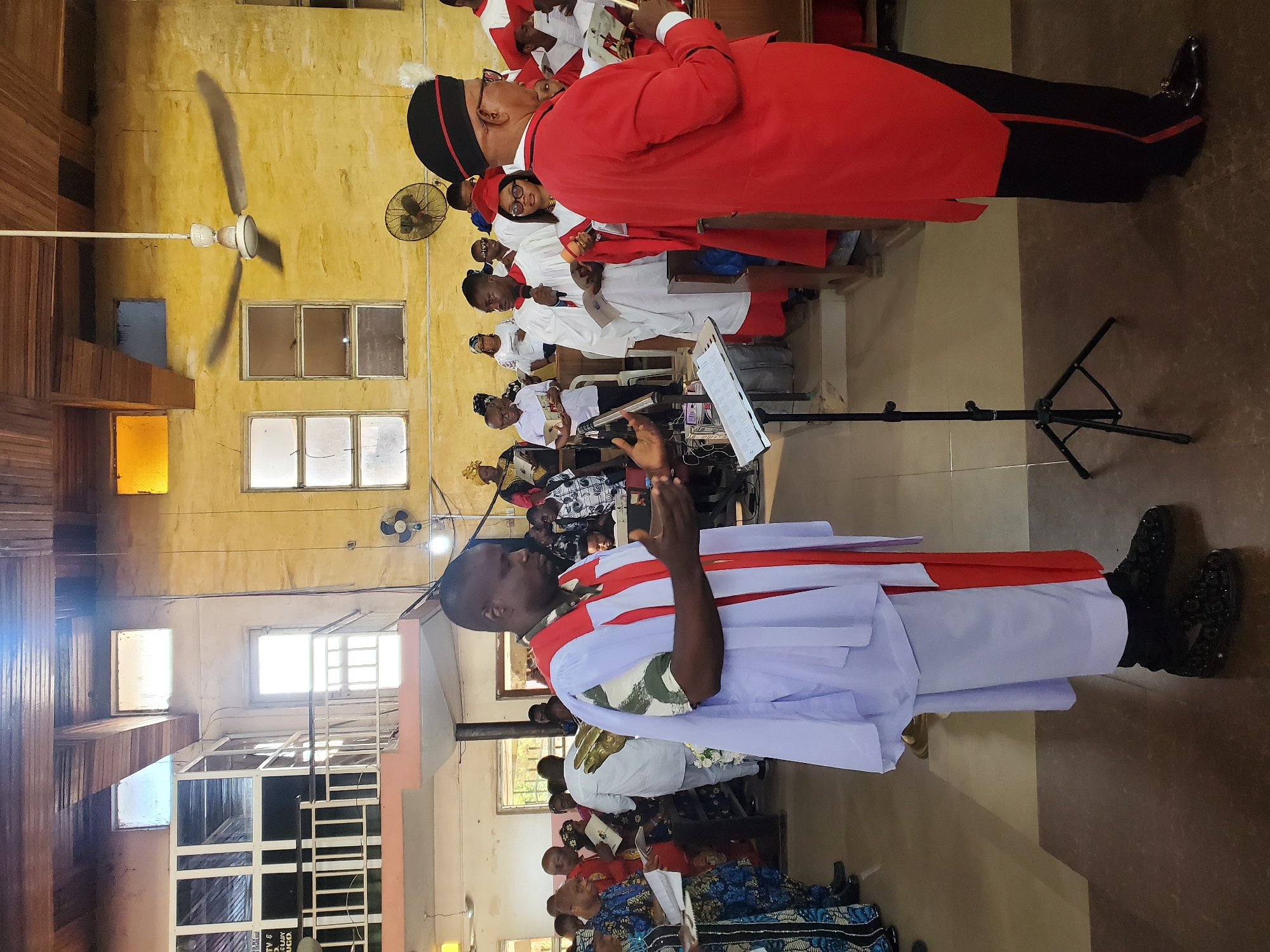 This image depicts an indoor religious gathering, likely a church service or choir performance. The scene is set in a large hall with light yellow, somewhat aged walls and wooden paneling on the left side. Natural light streams in through several rectangular windows. Ceiling fans are visible, including a large one in the upper left.
In the foreground, two men are prominent. On the right, a man wearing a red cassock with black trim and a black cap is seen from the side, looking towards the left (viewer's right). He wears glasses. To the left, another man in a white robe adorned with red and white stoles stands with his hands clasped or raised, possibly leading a prayer or song. He appears to be speaking into a microphone on a stand.
Behind these main figures, numerous individuals are arranged in rows, forming what appears to be a choir or congregational group. Many are dressed in white or red robes, while others wear vibrant, patterned traditional African attire. Some participants are holding books or music sheets. A music stand is visible in front of the group.
A small, dark sign in the background near the lower-left corner shows partially legible text including "GOSPEL," "ART," and "9 AM," suggesting a morning event with a gospel or religious arts focus. The overall atmosphere is one of focused participation and reverence.