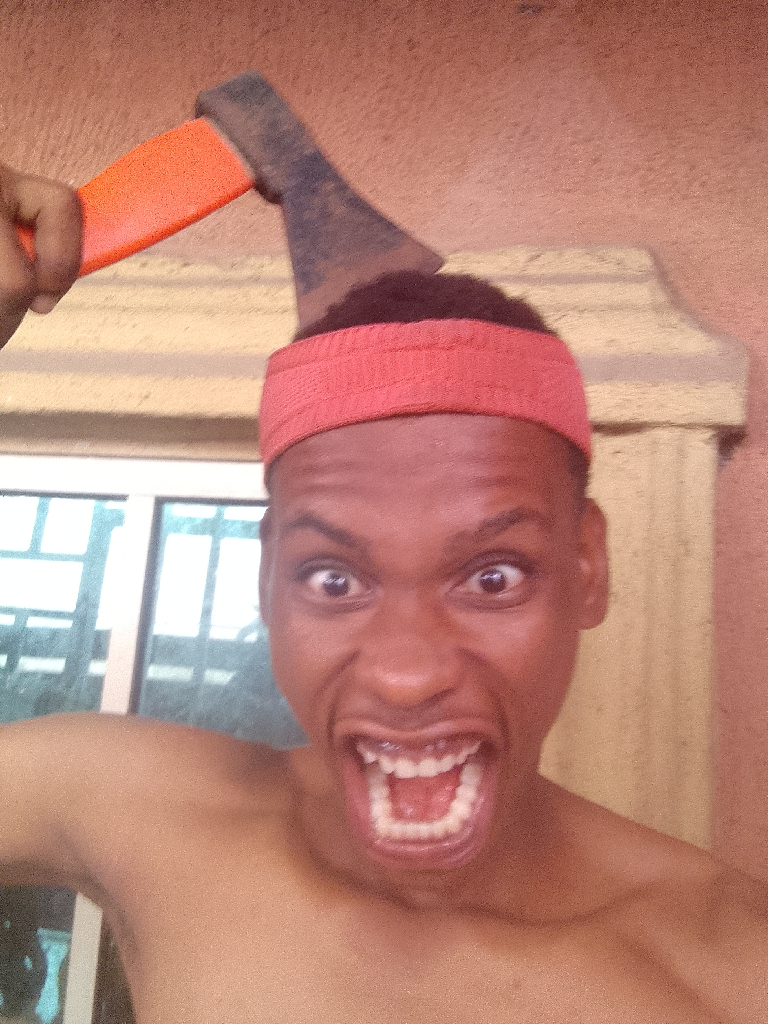 A topless man with a red headband is yelling and holding an ax. He is standing in front of a window. The location is Unknown, Unknown.