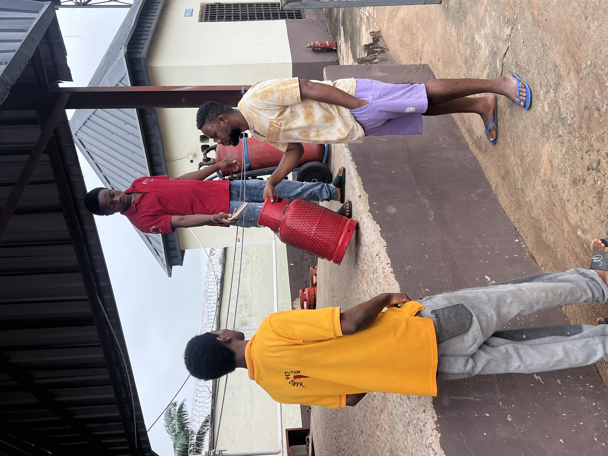 In Nkpor, Nigeria, three men are gathered outside a building with a corrugated metal roof. One man, wearing a red polo shirt and blue jeans, stands near a red gas cylinder and appears to be handing a lighter to another man who is holding the cylinder. This second man, wearing a tie-dye t-shirt and purple shorts, is looking at the first man. The third man, facing away from the camera and wearing a yellow polo shirt and grey jeans, is also present. Two other red gas cylinders are visible in the background. The scene takes place on a concrete surface next to a raised platform.