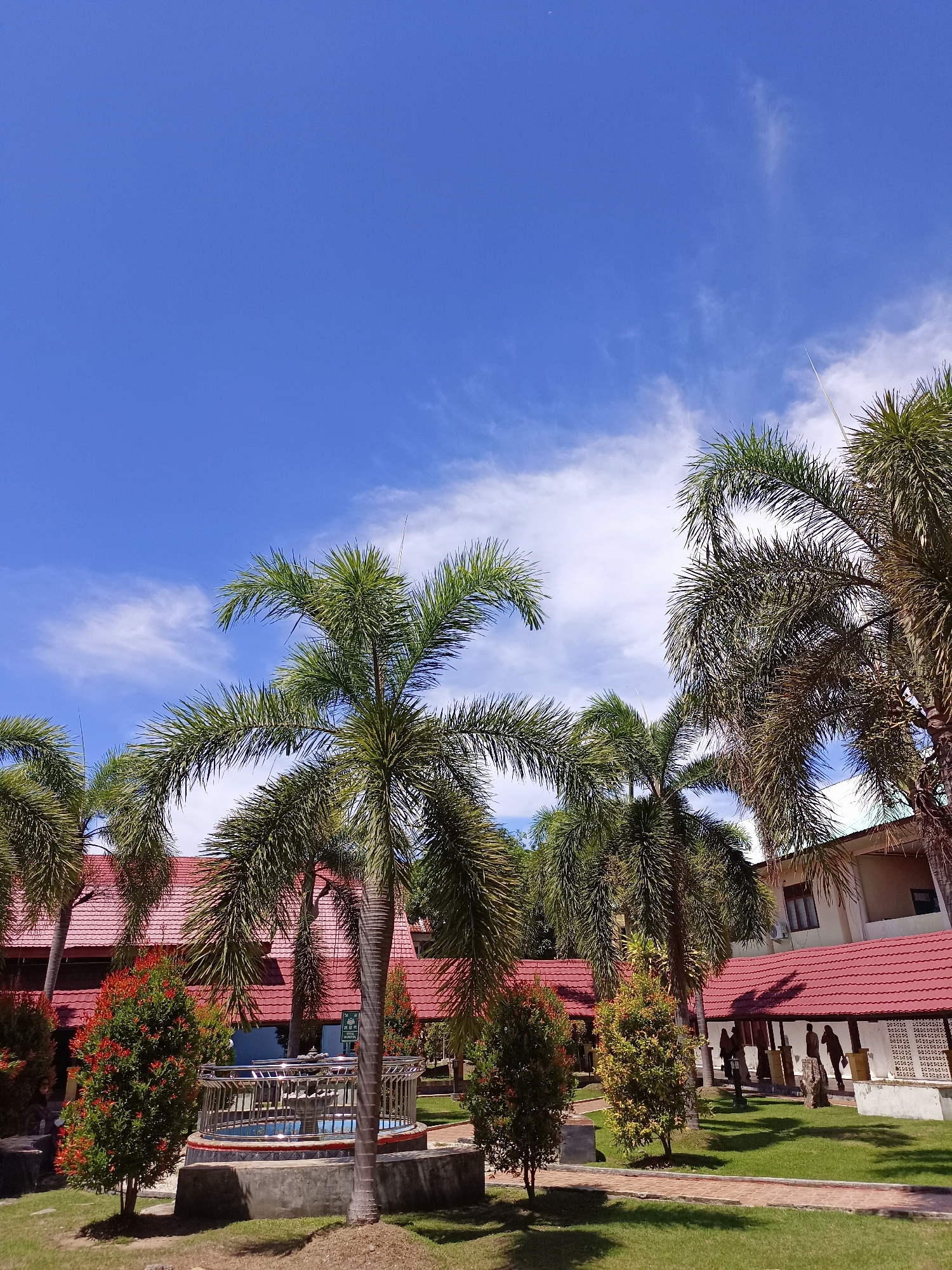 A daytime exterior shot shows a cluster of tall palm trees against a bright blue sky with scattered white clouds. In the foreground, a fountain with a metal railing is surrounded by lush green grass and some ornamental trees with red and green foliage. Behind the fountain, a building with a red tile roof and white walls is visible. The building appears to be part of a complex, with a covered walkway extending along its side. The scene suggests a tropical or subtropical environment, possibly a resort or a public park. The location is identified as Mesjid Reuntoh, Indonesia.