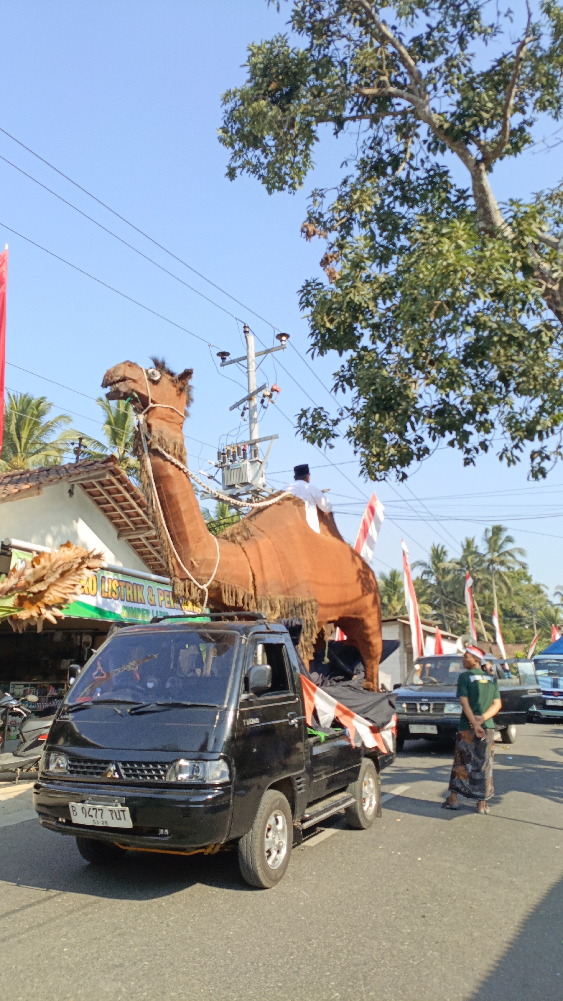 A replica of a camel, constructed from brown fabric and ropes, is being transported on the back of a black Mitsubishi pickup truck in Krajan Lor, Indonesia. The camel replica is large, extending over the top of the truck's cab and rear bed, and is adorned with decorative fringes and a rope halter. A person wearing a white shirt and a black peci hat is seated on the camel's back. The truck has a license plate that reads "B 9477 TUT." In the background, there are several Indonesian flags with red and white stripes, palm trees, buildings, and utility poles. A person in a green shirt and a batik sarong is standing on the right side of the road, next to another car. The scene appears to be part of a parade or a festive event, given the decorations and the presence of multiple vehicles and onlookers. The lighting suggests it is daytime with a clear blue sky.