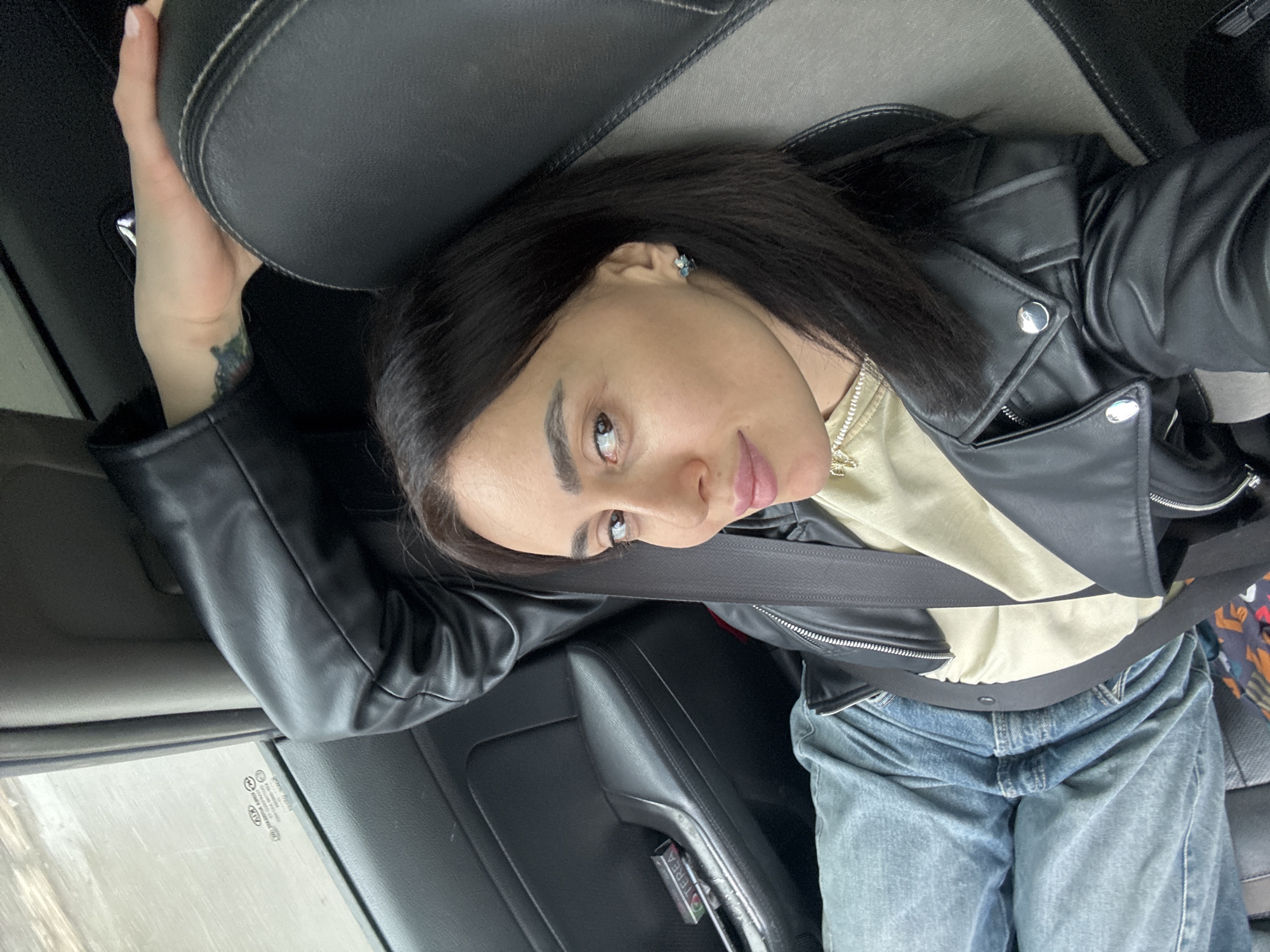 A single adult female is depicted in the passenger seat of a vehicle, in the city of Tbilisi, Georgia. She has dark hair, brown eyes, and is looking directly at the camera with a slight smile. She is wearing a black leather jacket over a light-colored (possibly beige or light green) top and blue denim jeans. A black seatbelt is fastened across her chest and lap. A delicate gold-colored chain necklace and small, multi-faceted earrings are visible. A small, dark tattoo is present on her left wrist. Her left hand rests on the top of the dark grey and black car seat headrest.
The vehicle's interior is predominantly dark, with a black door panel visible to the subject's left. On the door's armrest, a partially open rectangular pack of "TEREA" (an IQOS tobacco stick brand) is visible. Through the vehicle's window, which bears a "KIA MOTORS" decal, a blurred, indistinct outdoor environment is visible, suggesting the vehicle is in motion or the background is out of focus.