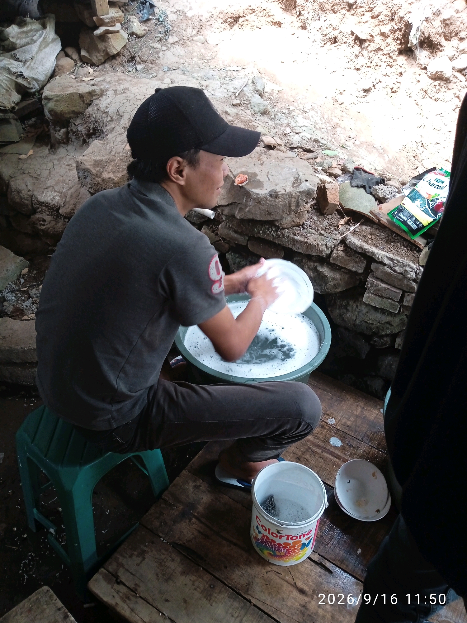 A young man wearing a black baseball cap and a dark gray polo shirt is sitting on a green plastic stool and washing dishes in a large, light green basin filled with soapy water. The basin is situated on a wooden surface. Next to the basin, there is a white bucket with the "ColorTone" logo visible, and an empty white bowl. In the background, there is a rough stone wall and some loose rocks, suggesting an outdoor or unfinished setting. A portion of a black garment is visible in the right foreground, obscuring part of the scene. A packet with green and white packaging, possibly containing food or spices, is on the ground near the stone wall. The image is timestamped 2026/9/16 11:50. The overall environment appears rustic and possibly rural.