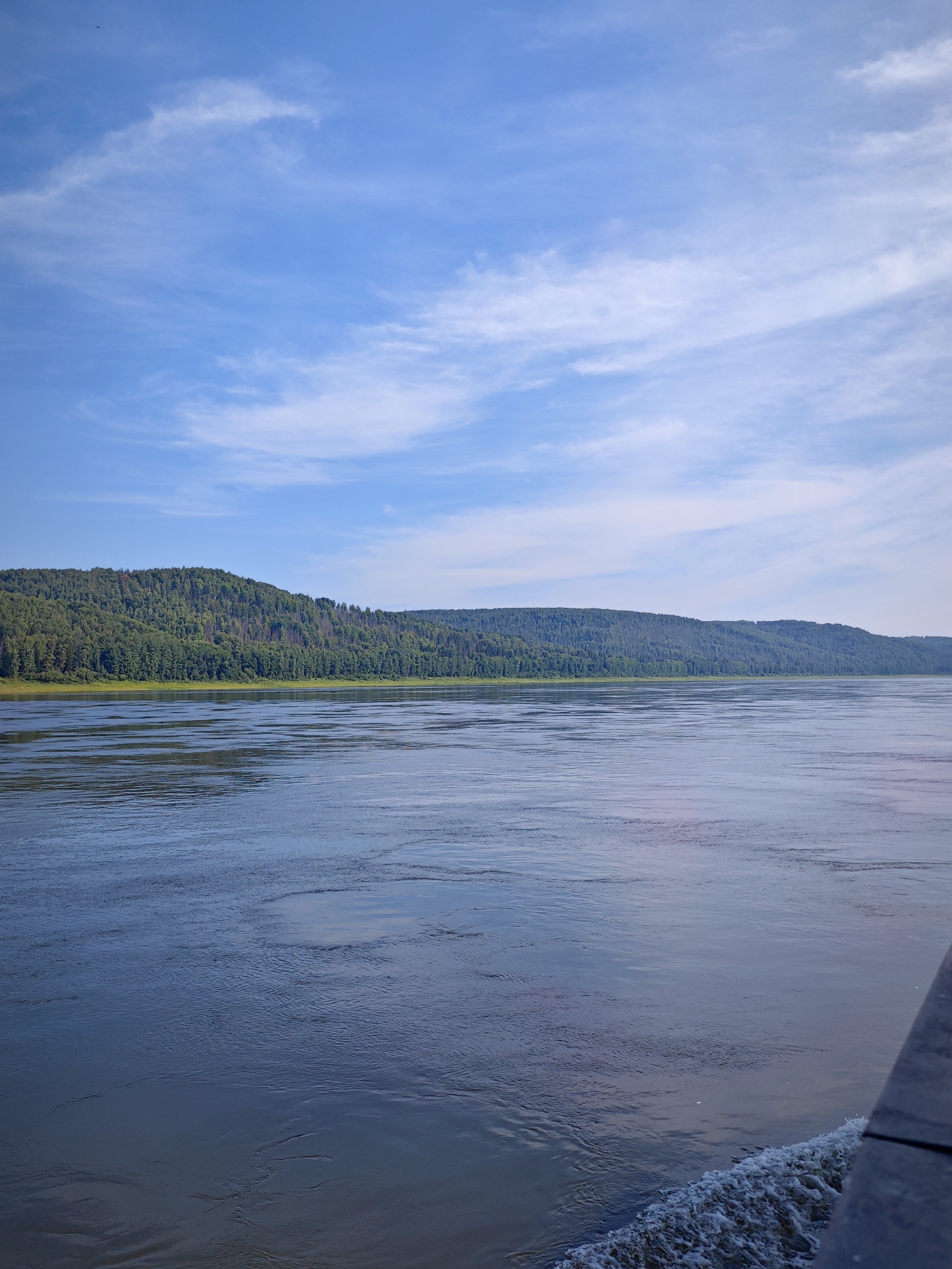 A wide, calm river stretches across the foreground of the image, reflecting the blue sky and wispy white clouds. On the far bank, a dense, green forest covers rolling hills that recede into the distance. The sky is a bright, clear blue with streaks of thin clouds. The water's surface is mostly smooth, with subtle ripples and reflections. A dark, wooden structure, possibly part of a boat or dock, is visible in the lower right corner of the frame, with a wake of white foam trailing behind it in the water. The scene depicts a natural landscape under a bright sky.