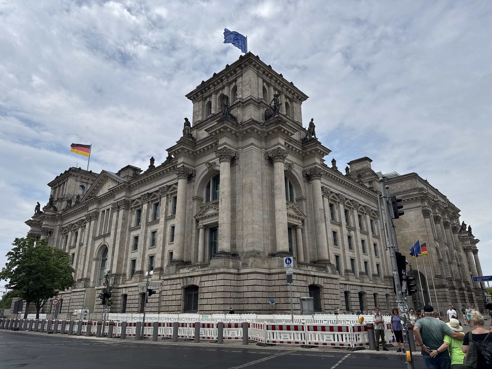 The image shows the Reichstag building in Berlin, Germany, a historic landmark and seat of the German Parliament. The building is constructed of stone and features classical architecture with columns and ornate detailing. Two flags are visible: the German flag and the flag of the European Union, flying from the building's towers.
In the foreground, a street is lined with temporary barriers, suggesting ongoing construction or security measures. Several people are seen walking on the sidewalk and across the street, some of them tourists observing the building. The sky is cloudy, indicating an overcast day. Traffic lights and street signs are also present, indicating a busy urban environment.
The overall scene depicts a prominent government building in Berlin, with elements of tourism and urban activity surrounding it. The presence of both national and supranational flags highlights the building's significance in the context of both Germany and the European Union.