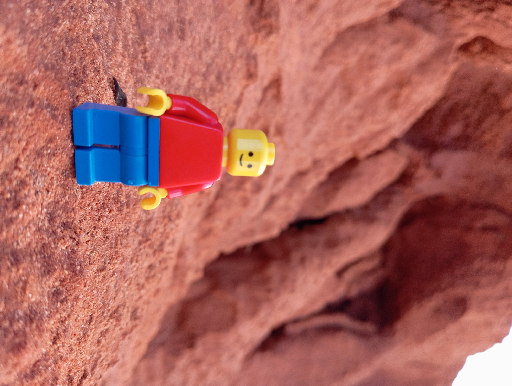 This image presents a close-up view of a standard Lego minifigure set against a rugged, textured, reddish-brown rock face. The minifigure, rendered in vibrant primary colors, features a yellow head with a cheerful, printed smiling face, a bright red torso, and blue legs, with yellow hands completing its classic look.
The minifigure is positioned vertically on the granular surface of the rock, which exhibits a coarse, earthy texture with varying shades of reddish-brown, hinting at a sandy or sandstone composition. The background, largely out of focus, continues the reddish-brown motif, depicting blurry geological formations that suggest a desert-like or canyon environment. Deeper, shadowed areas and recesses are visible in the background, indicating crevices or more complex rock structures.
The scene is illuminated by natural daylight, providing soft lighting that highlights the intricate texture of the rock and the smooth plastic of the Lego figure. The location context, Mui Ne, Vietnam, suggests these formations are part of the region's famous red sand dunes or fairy stream geology. The minifigure’s smiling expression adds a whimsical and positive tone to the otherwise stark natural setting. There is no visible text in the image.