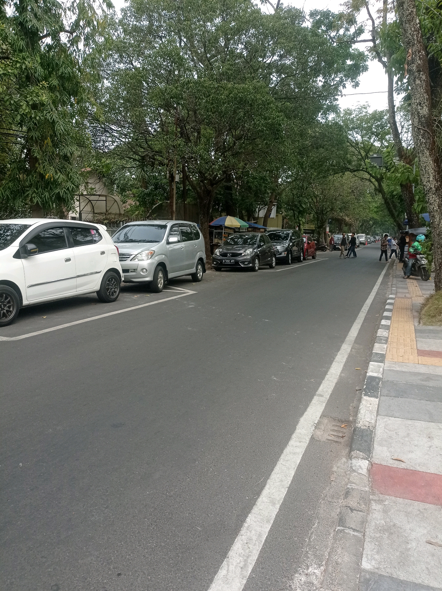 The image depicts a street scene in Bandung City, Indonesia, with a focus on parked cars and pedestrian activity. A white hatchback and a silver minivan are prominently parked on the left side of the asphalt road, within marked parking bays. Further down the street, several other cars are also parked, including a dark grey hatchback and a red car.
On the right side of the road, a sidewalk is visible, paved with different colored tiles, including yellow and red. Several individuals are walking on the sidewalk and crossing the street in the distance. A person on a scooter is parked on the right edge of the frame.
The environment is characterized by lush green trees lining the street, suggesting a well-maintained urban area. The weather appears overcast, with a generally muted natural light. The presence of power lines overhead indicates an urban infrastructure. The scene is a snapshot of everyday life on a city street, capturing a moment of movement and stillness.