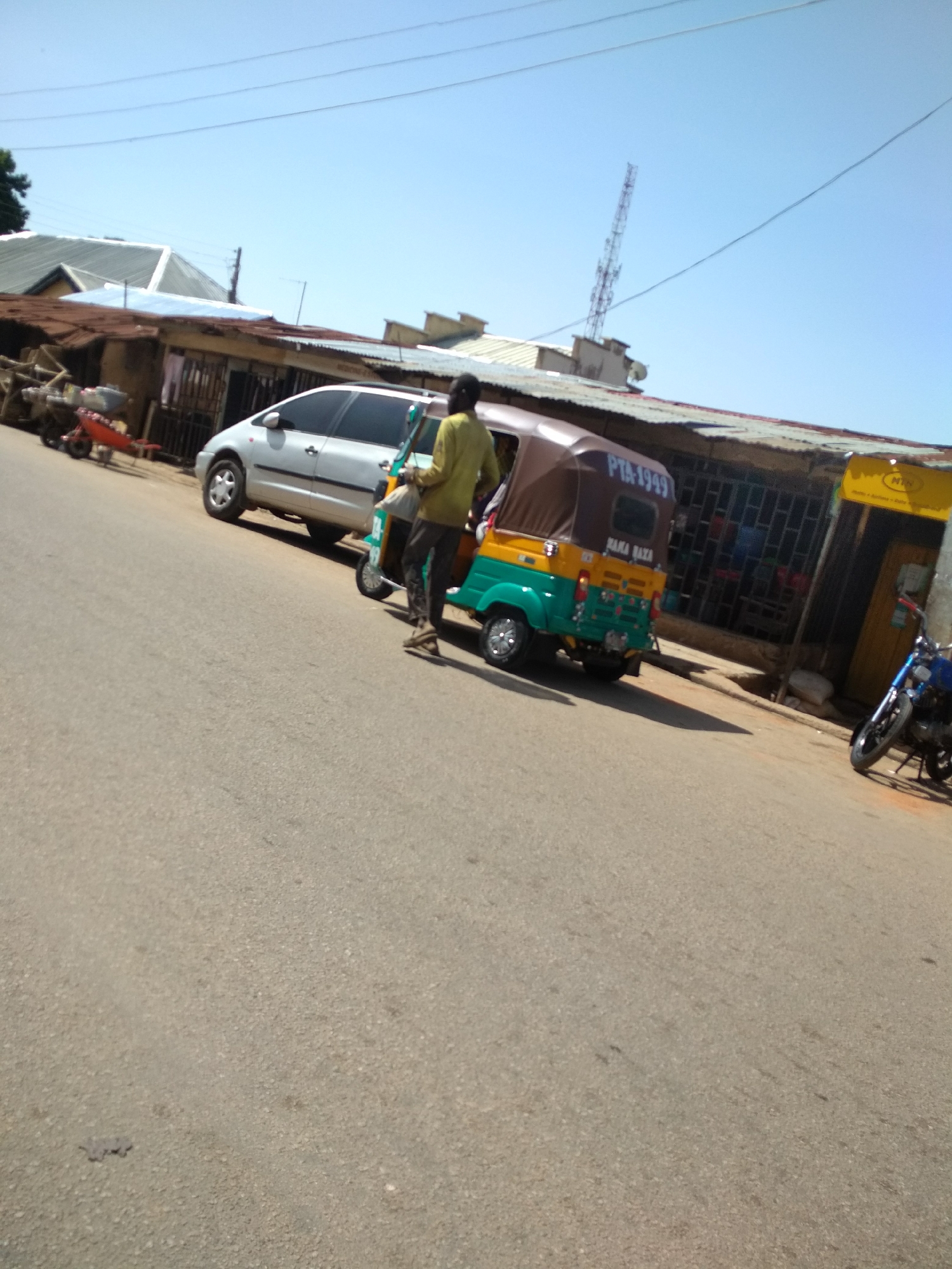 This image depicts a bustling street scene under a clear, bright blue sky, suggesting mid-morning or afternoon. The paved road slopes diagonally across the frame.
In the foreground, a person wearing a yellow-green long-sleeved shirt and dark pants stands beside a green and yellow three-wheeled auto-rickshaw or tricycle. Visible on the rear of the tricycle are the identifiers "PTA 1949" and "RAMA BABA". Parked behind the tricycle is a light-colored (possibly silver or white) minivan or car. Further down the street on the left, a red handcart or wheelbarrow is present.
The background features a row of buildings, characterized by their corrugated metal roofs, some showing signs of rust. One building on the right side of the road has a yellow awning or sign with the prominent text "MTN" clearly visible, along with some smaller, illegible text below it. A blue motorcycle is partially visible, parked near this building. High above the buildings, a tall, slender communication tower (likely a cell tower) stands against the sky, accompanied by overhead power lines.
The scene captures typical street activity in what appears to be an urban or semi-urban environment. The vehicles, architecture, and "MTN" branding suggest a location possibly in Africa.