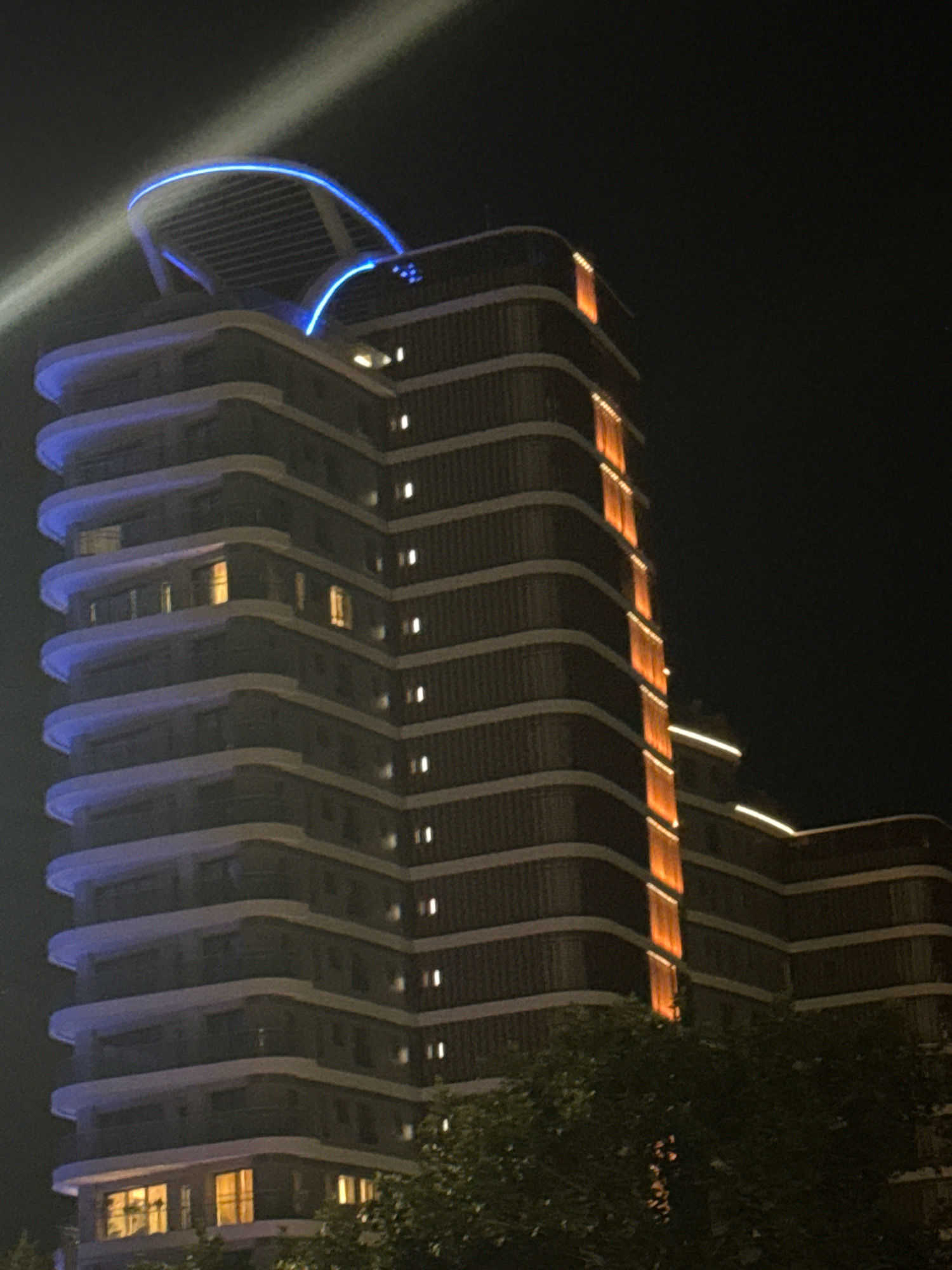 An exterior night shot features a tall, modern building with a curved facade. The building is illuminated by a combination of blue and orange lights. Blue lights outline the curved balconies and a distinctive architectural feature at the top of the building, which appears to be a large, illuminated arch. Vertical orange lights highlight sections of the building's facade, creating a striped effect. Small, white lights, likely from windows, are visible on various floors. The lower portion of the image shows the dark foliage of trees, partially obscuring the base of the building. A bright white light beam cuts across the upper left corner of the image, originating from outside the frame. The building is located in Germasogeia Municipality, Cyprus.