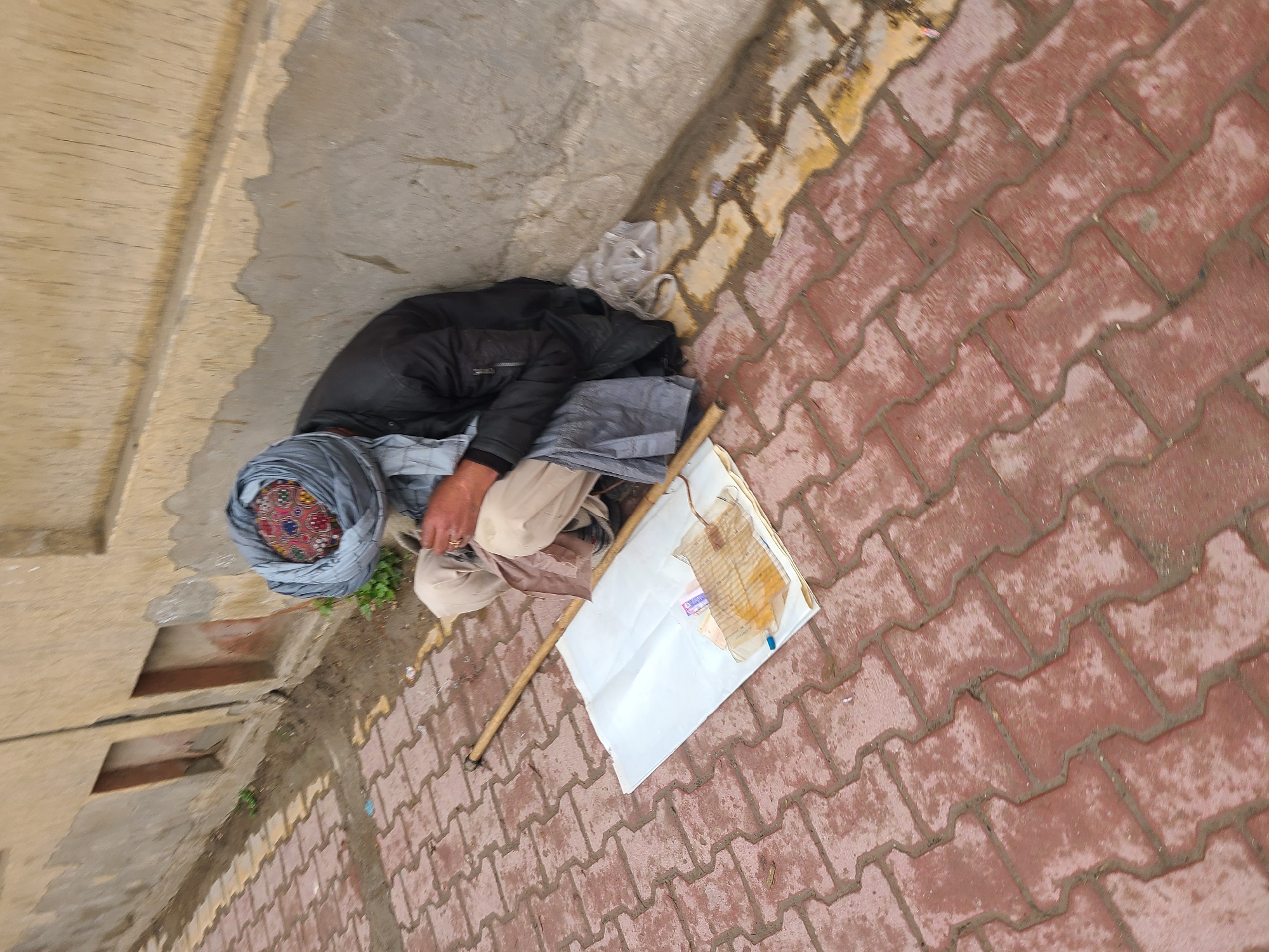 The image depicts a man sitting on the ground in Quetta City Tehsil, Pakistan. He wears a dark jacket, light tan pants, and a colorful patterned turban. He is positioned near a concrete wall and a red brick-paved area. Next to him is a white paper sign held up by a stick.