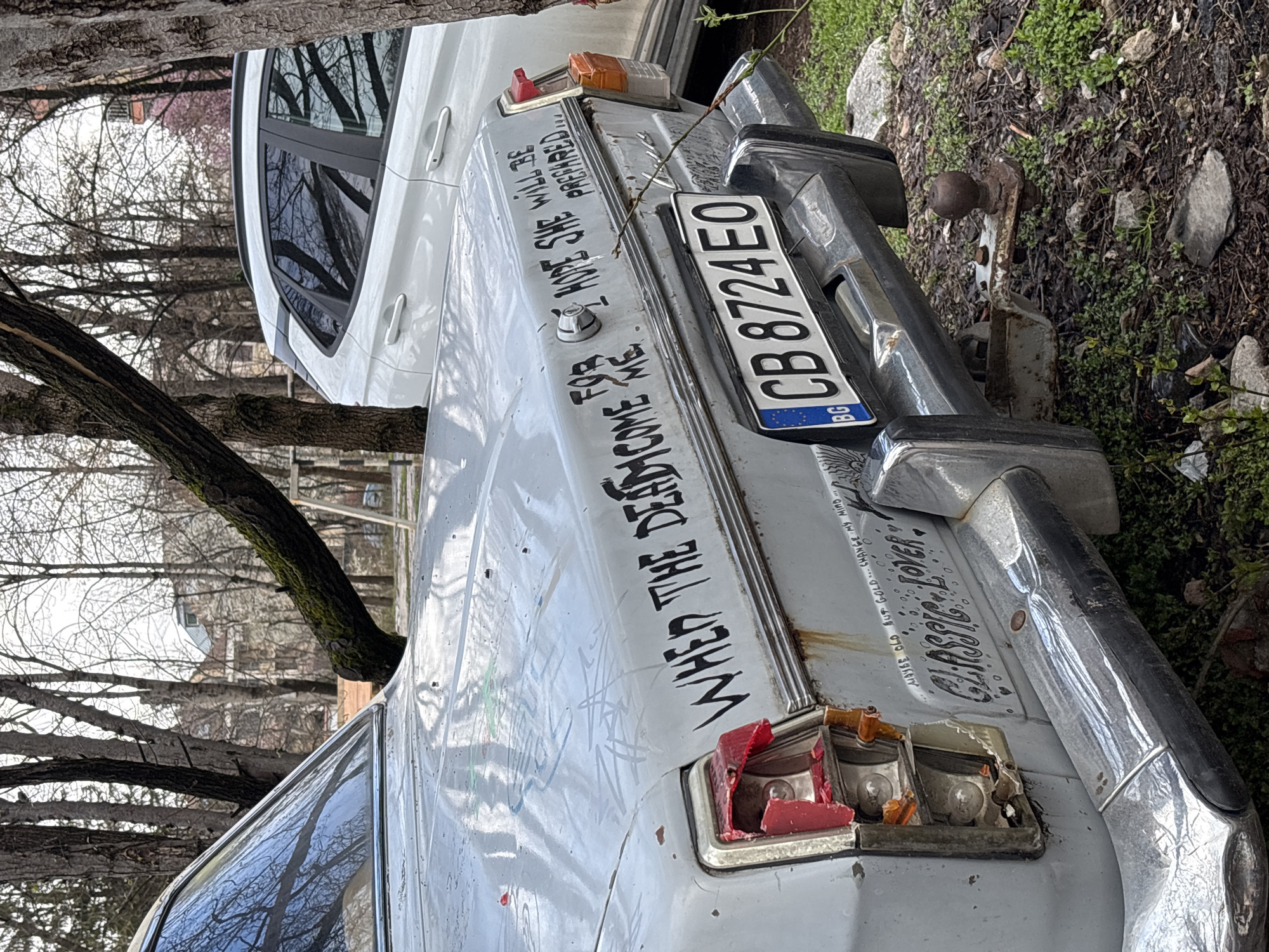 The image shows the rear of a faded white car, possibly a Lada, parked outdoors with a damaged right tail light. The license plate reads "CB 8724 EO," indicating it's from Bulgaria. Graffiti is scrawled on the trunk, including "WHEN THE DEAD COME FOR ME HOPE SHE WILL BE PREPARED...". Other phrases like "Classic Lover" and "Maybe old but gold..change my mind..." are also visible. The background shows bare trees, indicating it's likely late fall or early spring. The car is parked on ground covered with vegetation and rocks. The location is probably a neglected area within the city of Sofia, Bulgaria, as indicated by the license plate.