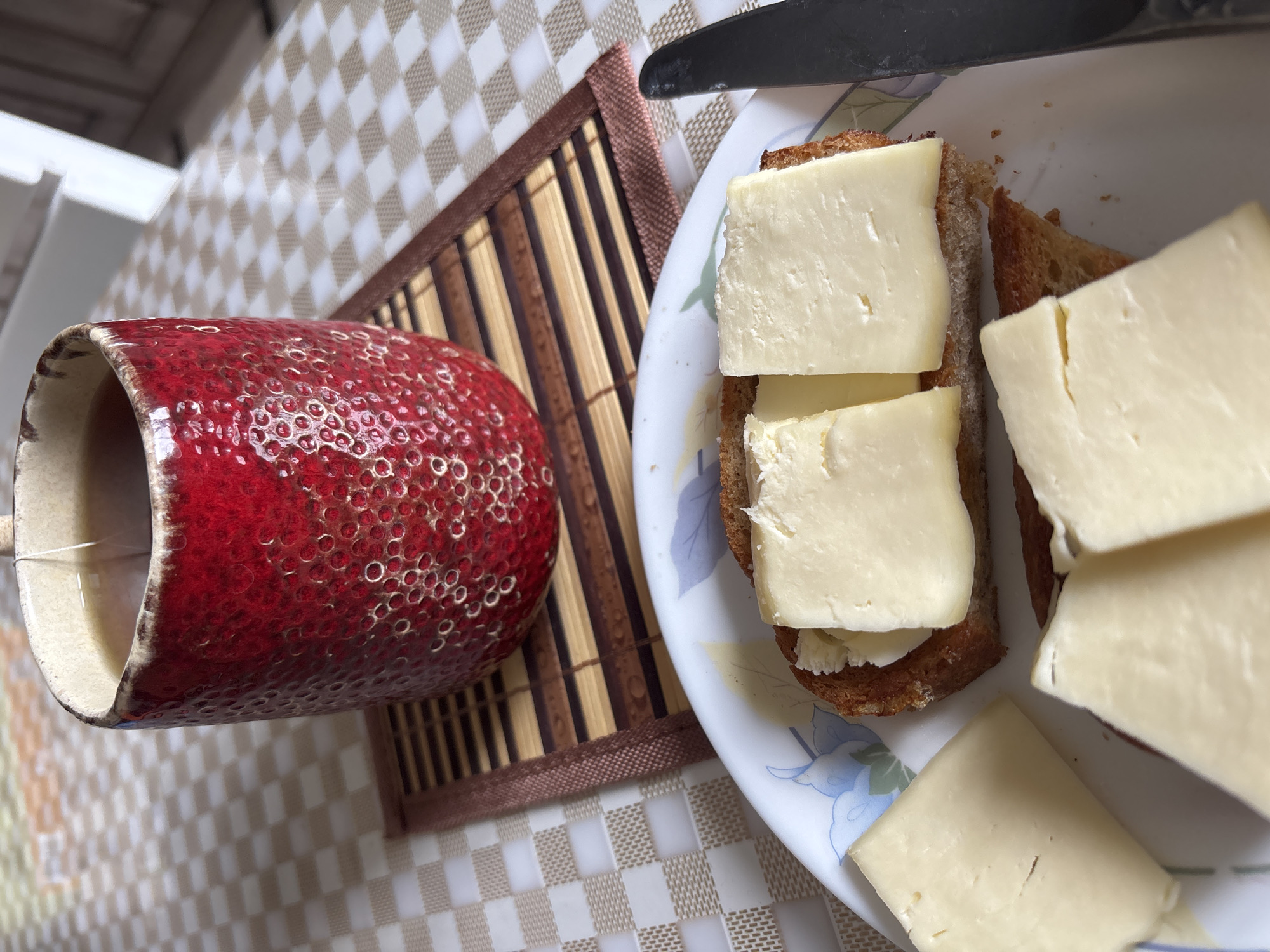 A mug of tea sits on a checkered tablecloth next to a plate of open-faced sandwiches topped with cheese. The mug is red with a textured surface and a tea bag string hanging out. The sandwiches are on toasted bread with slices of pale yellow cheese. A black knife is partially visible on the right side of the plate. The background is blurred, showing part of a chair and suggesting an indoor setting. There is no visible indication of the location being in Bila Tserkva, Ukraine.